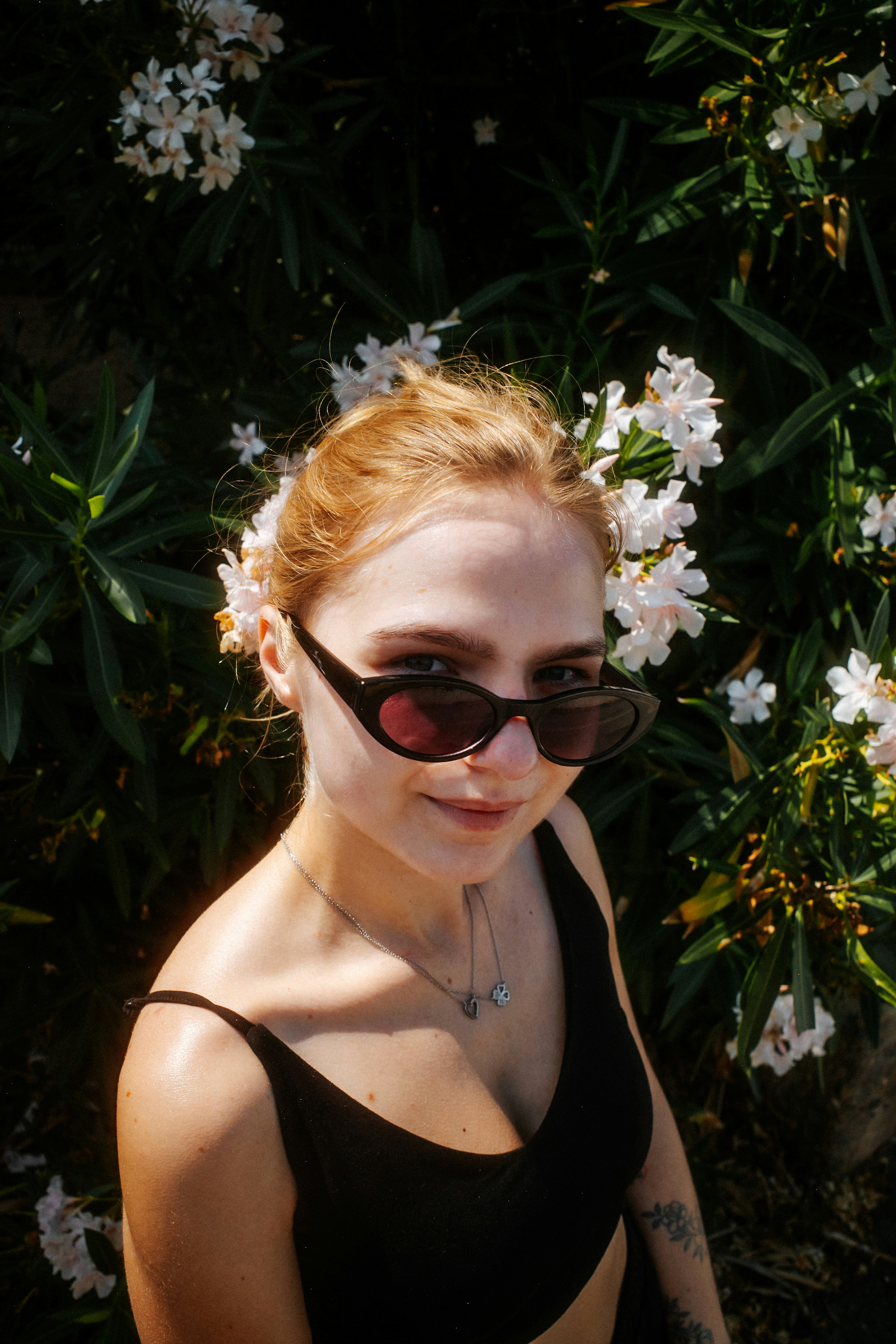 Une femme aux cheveux blonds, portant des lunettes de soleil et un haut noir devant des fleurs blanches.