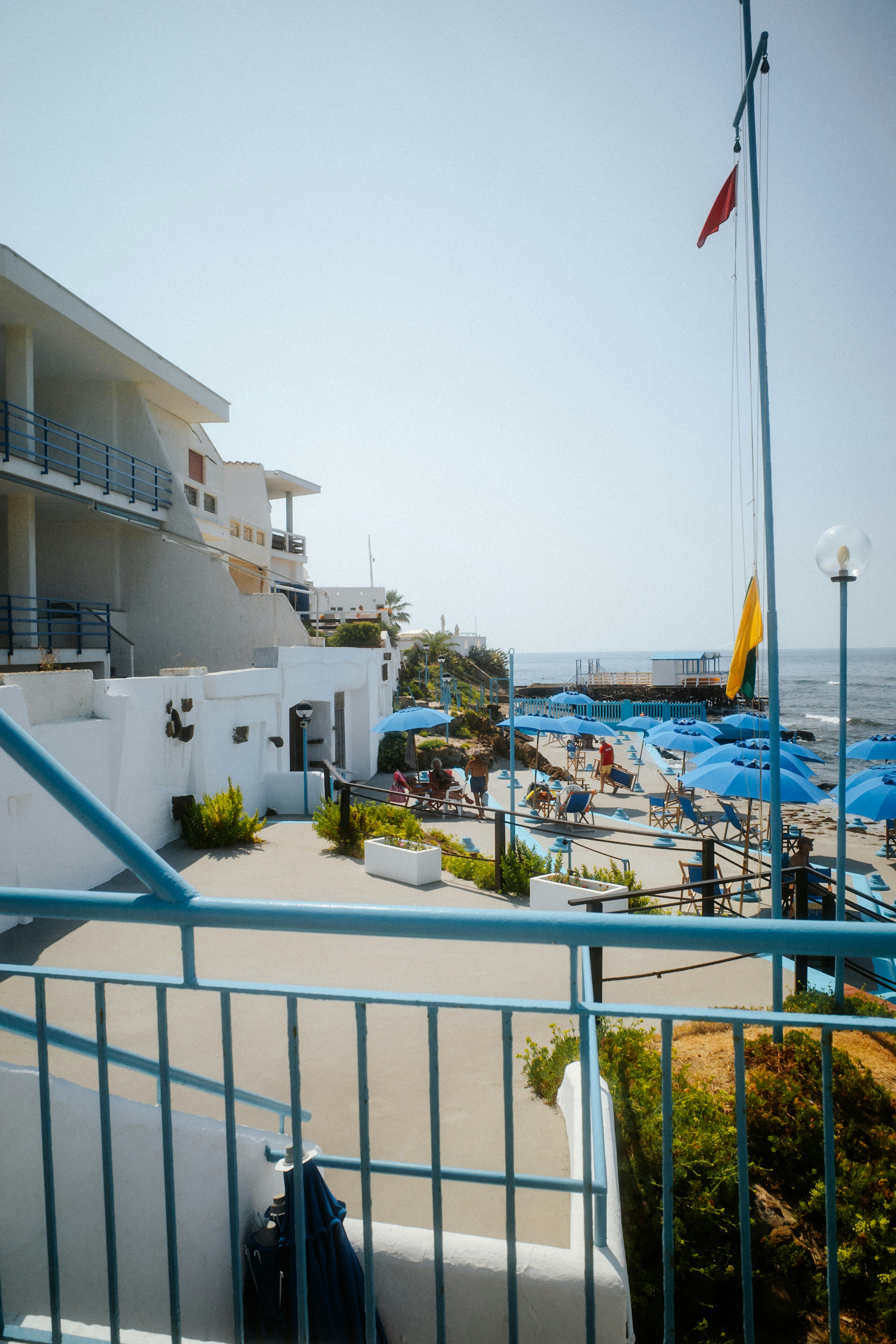 A white hotel terrace overlooking a beach with blue umbrellas and people.
