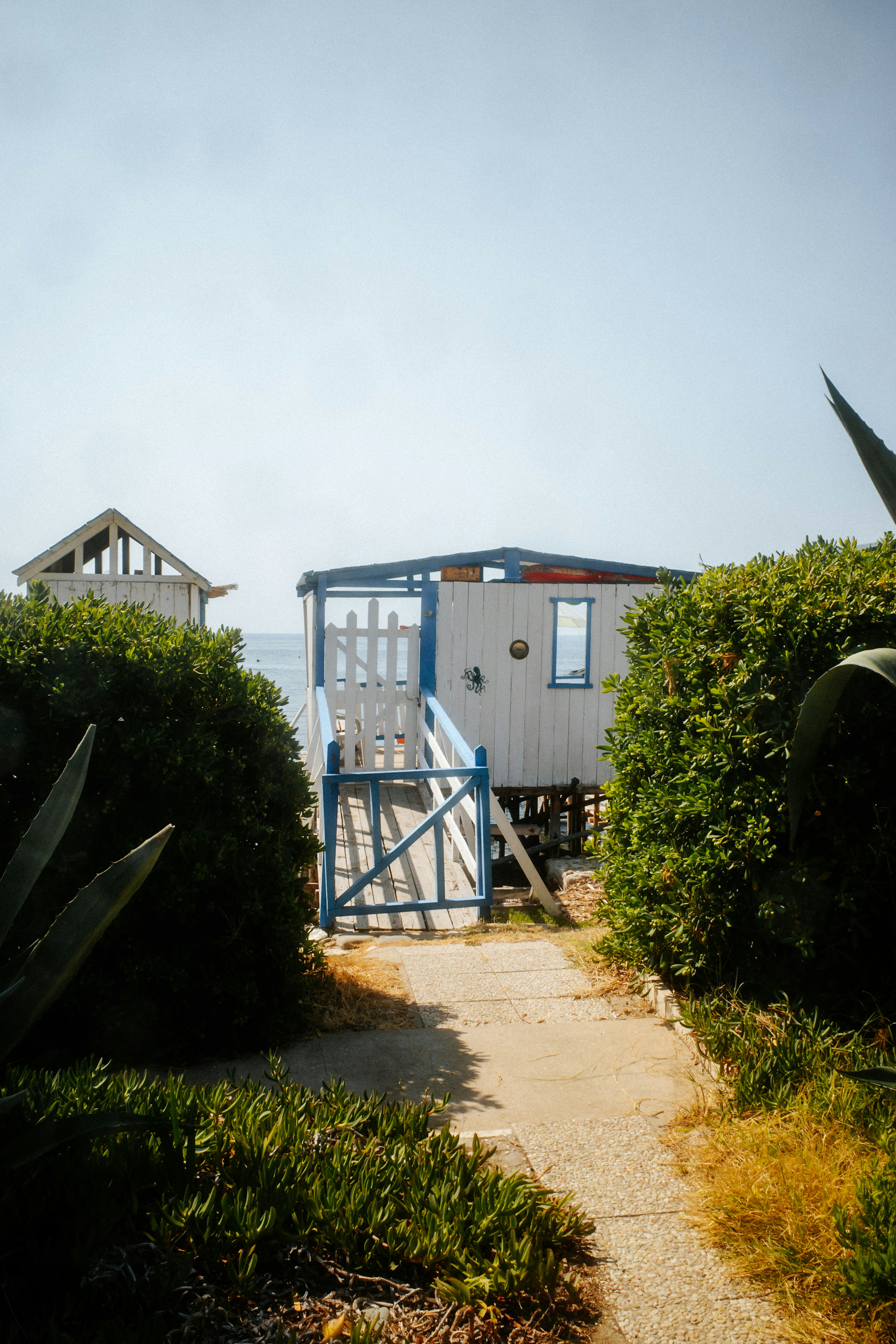 A white wooden beach cabin with blue trim at the end of a path.