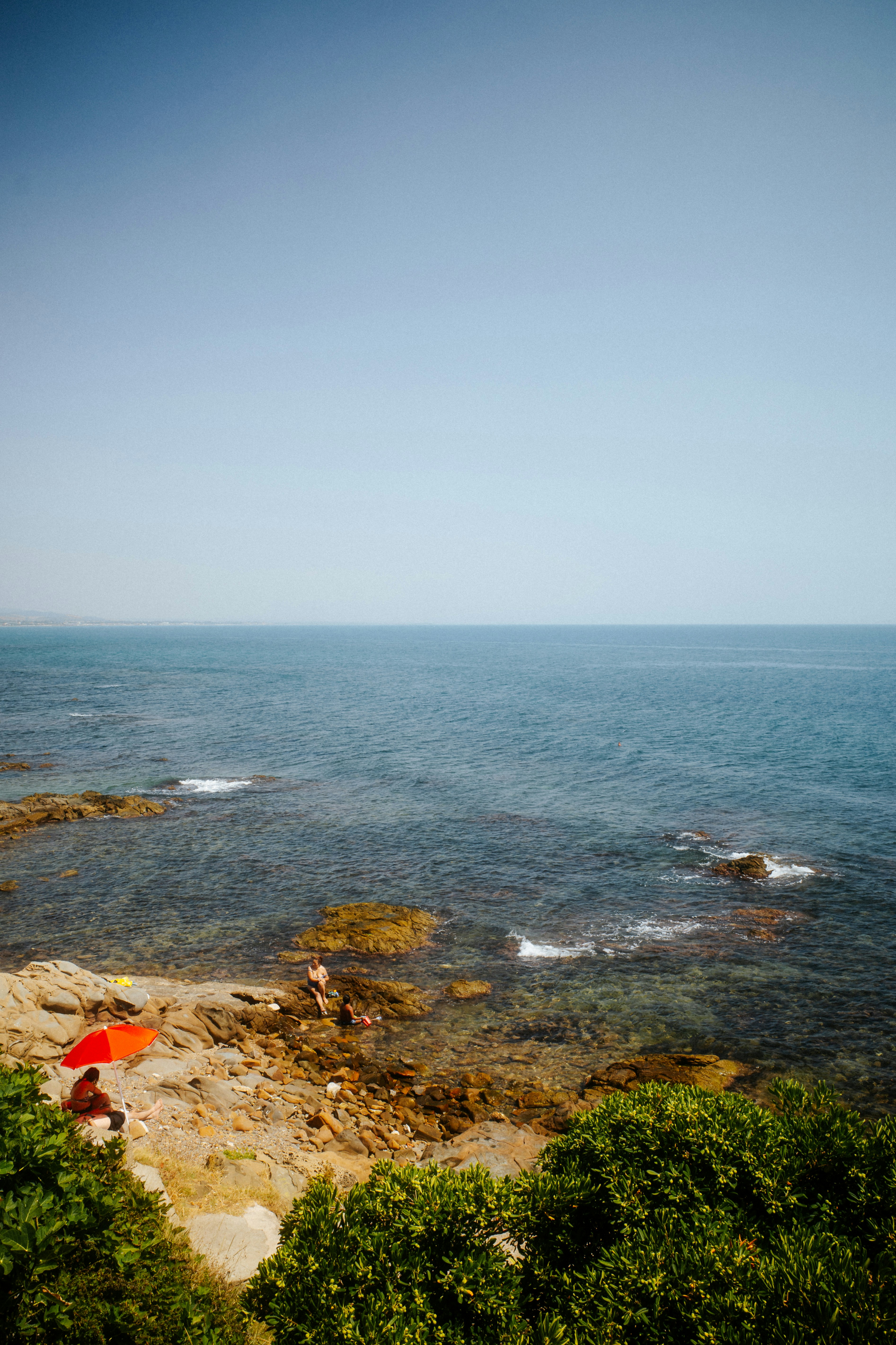 People on a rocky beach with a red umbrella by the blue sea.