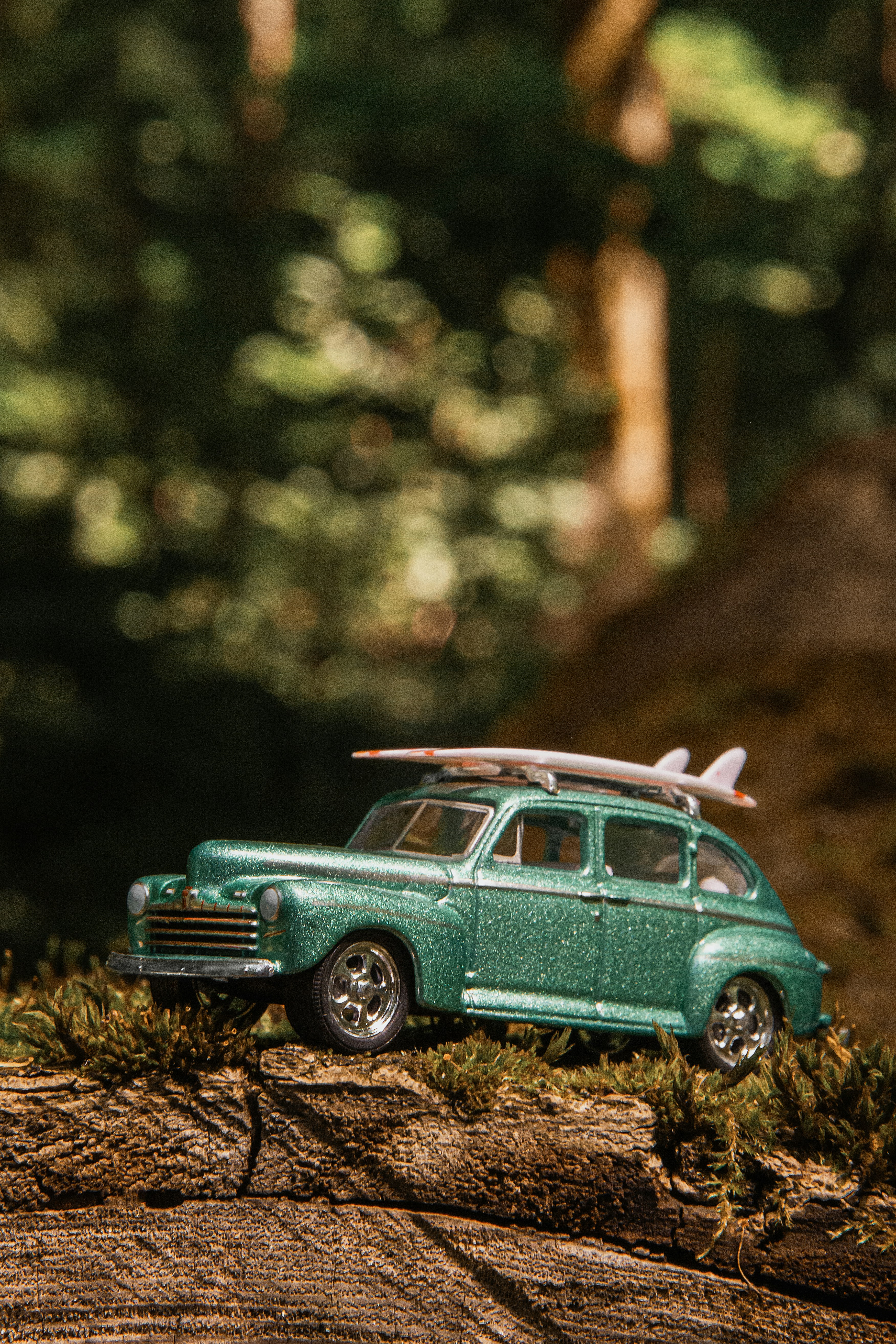 A green toy car with a surfboard on its roof on a mossy log.