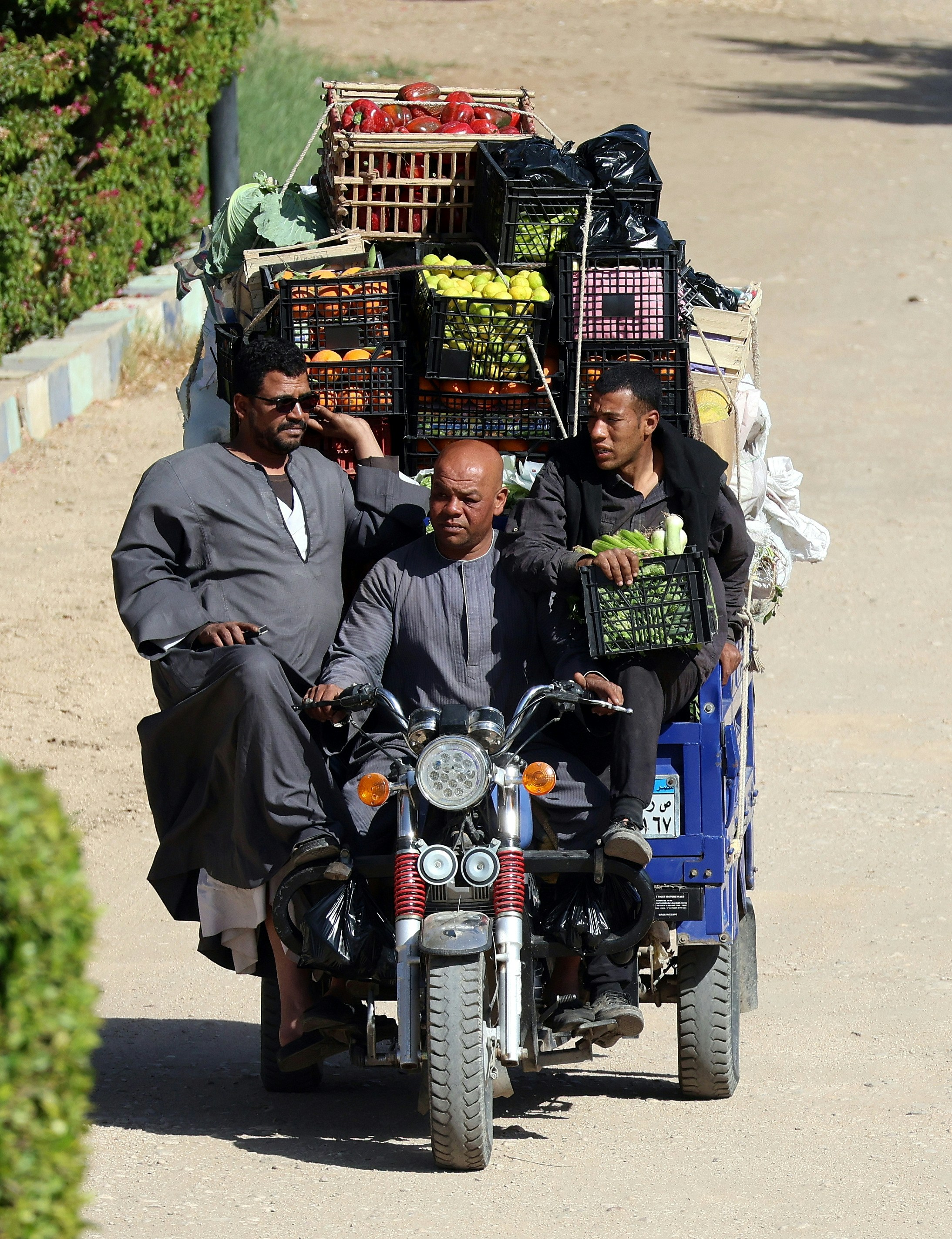 Three men riding a blue motorized tricycle loaded with crates of fresh produce.