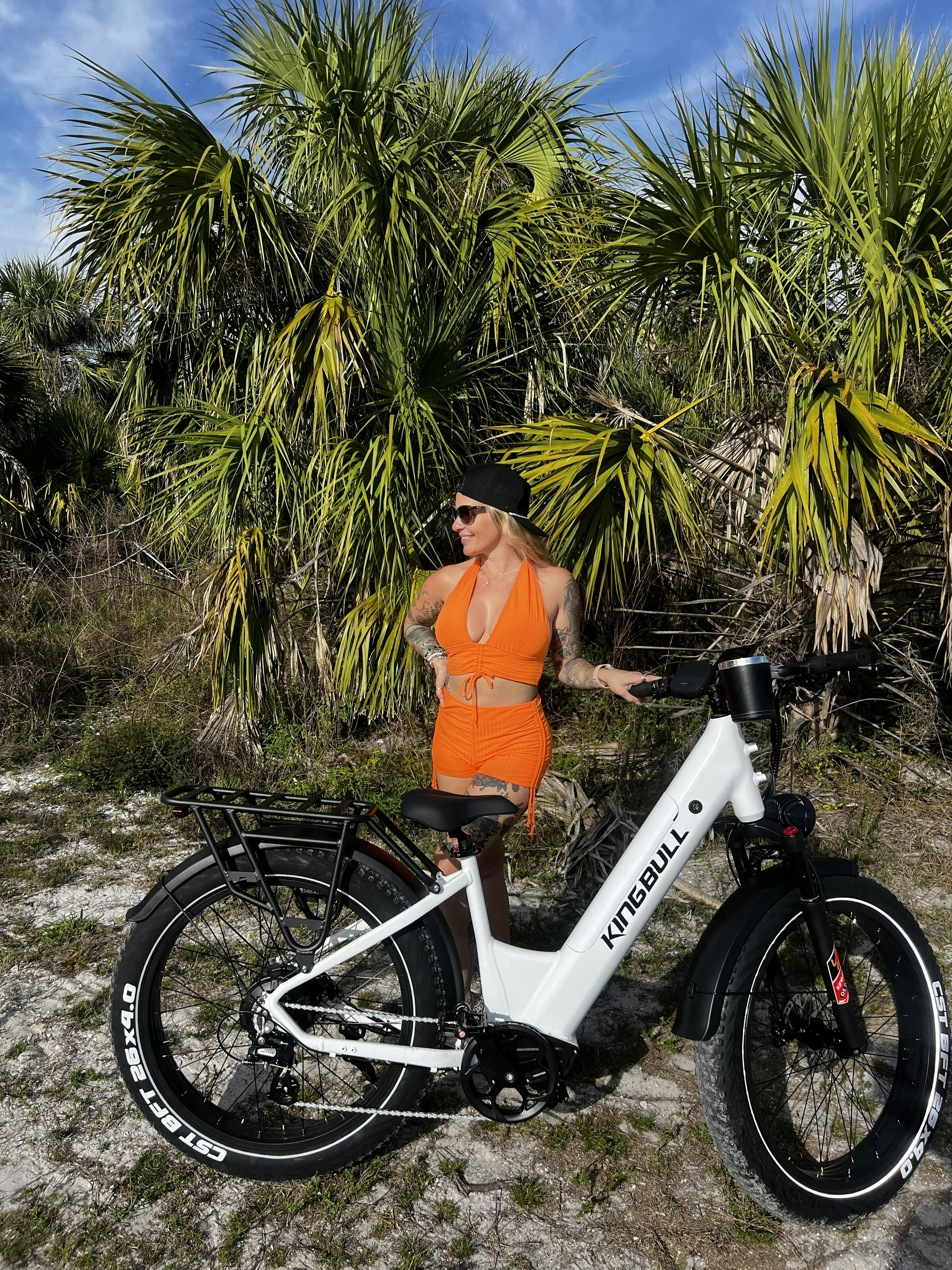オレンジ色の服を着た女性が、ヤシの木のそばで白い電動自転車のそばに立っている