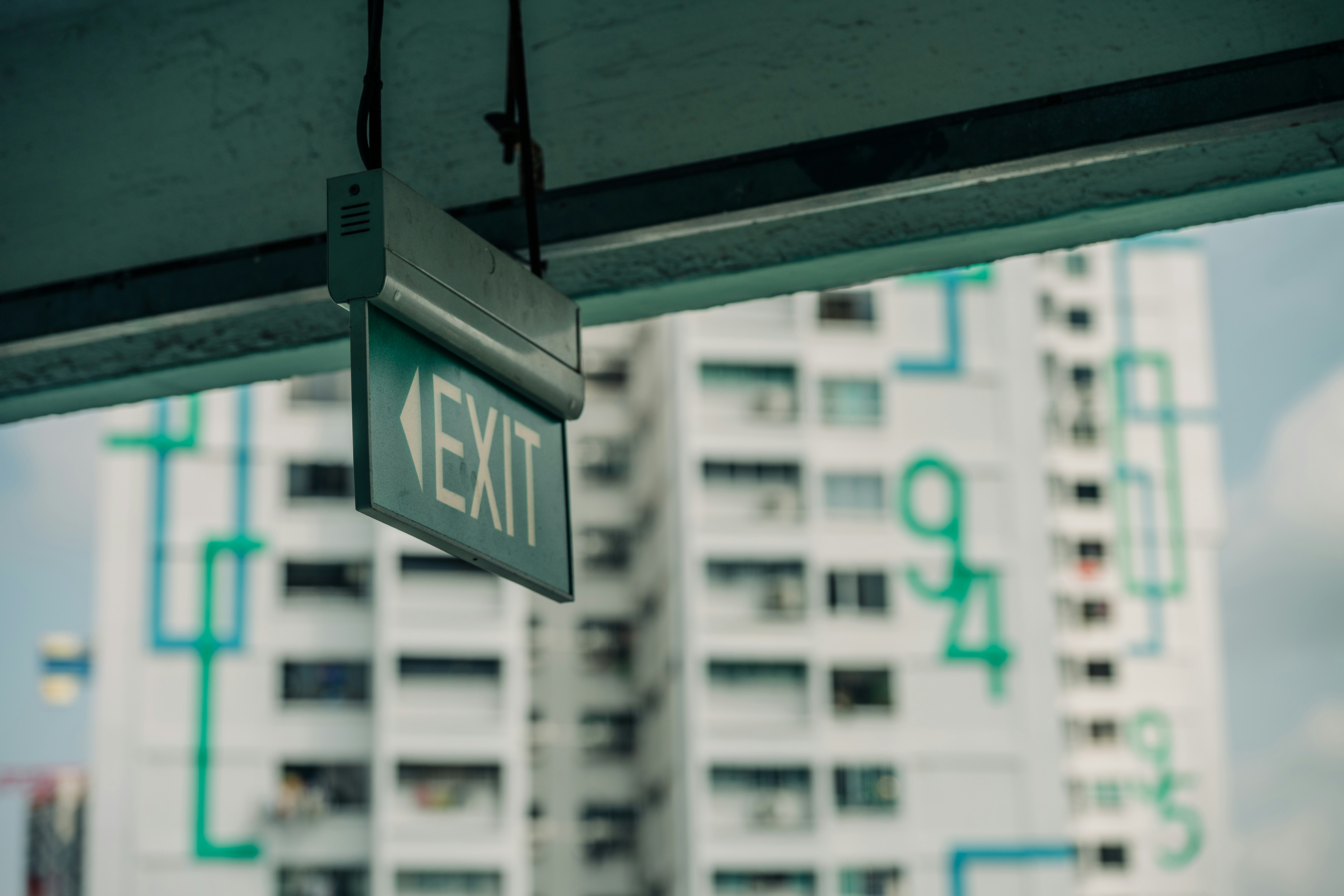 A green exit sign hanging from a ceiling before a tall apartment building.