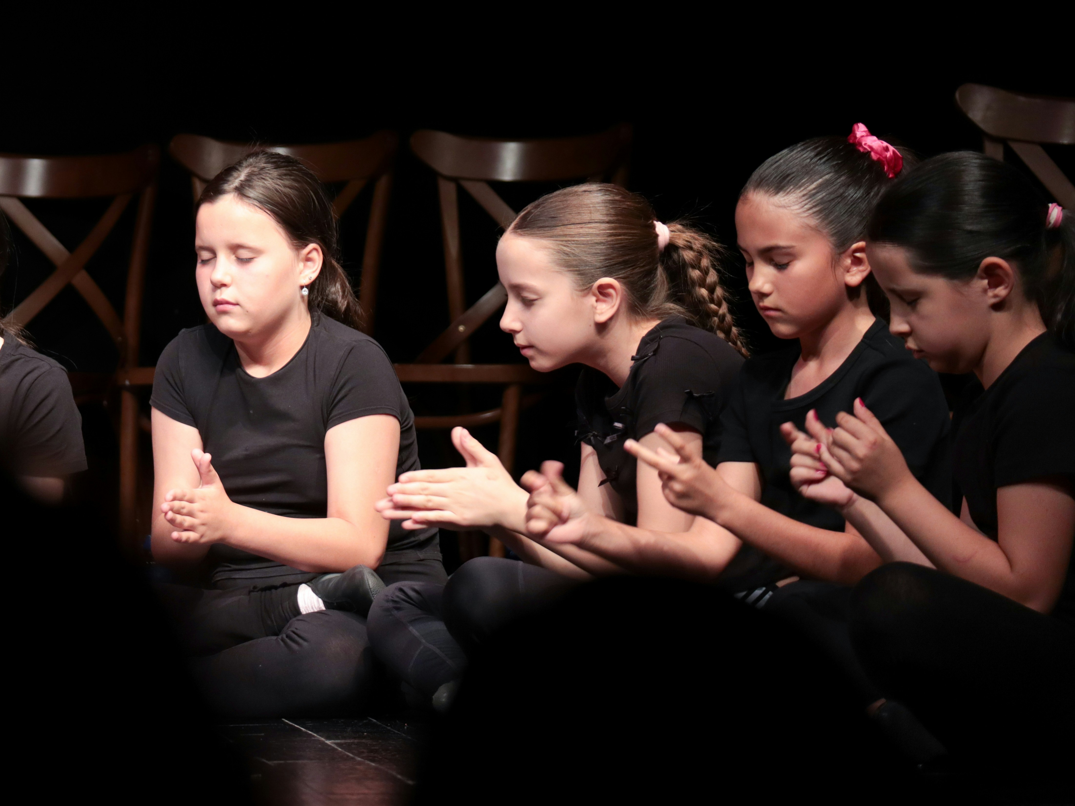 Four children in black clothing sitting on a stage with their hands together