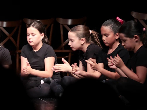 Four children in black clothing sitting on a stage with their hands together.