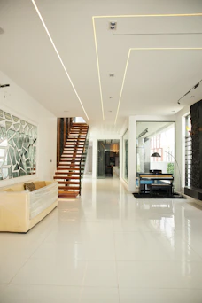 A modern living room with a wooden staircase, a piano, and white tiled floors.