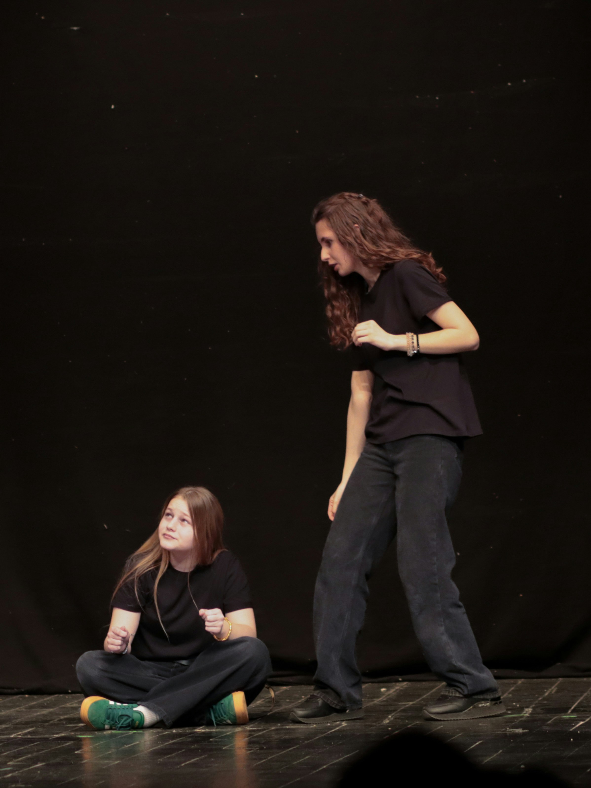 A person sitting on a stage floor looking up at another person standing