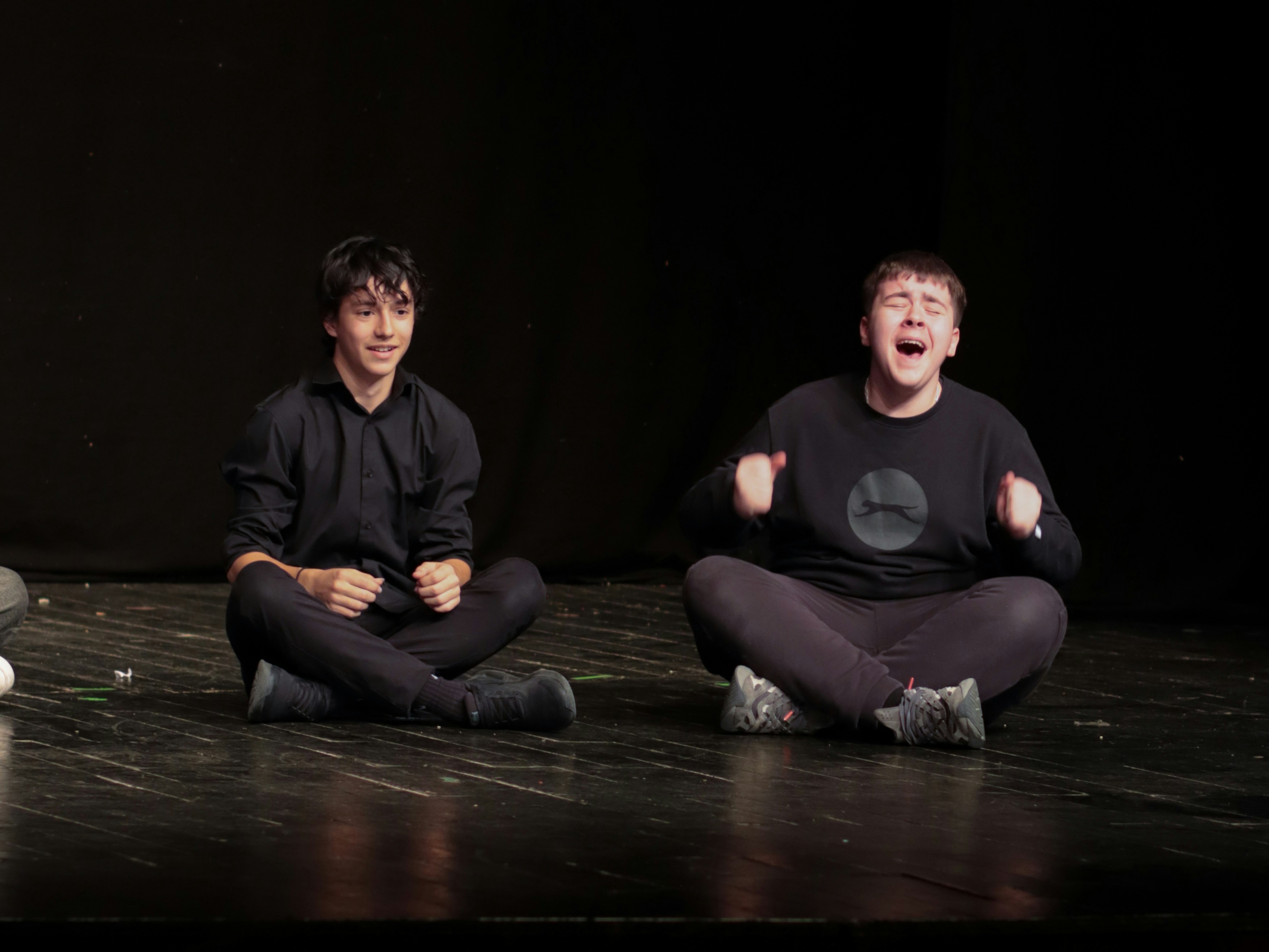 Two people sitting cross-legged on a dark stage, one laughing with raised fists.