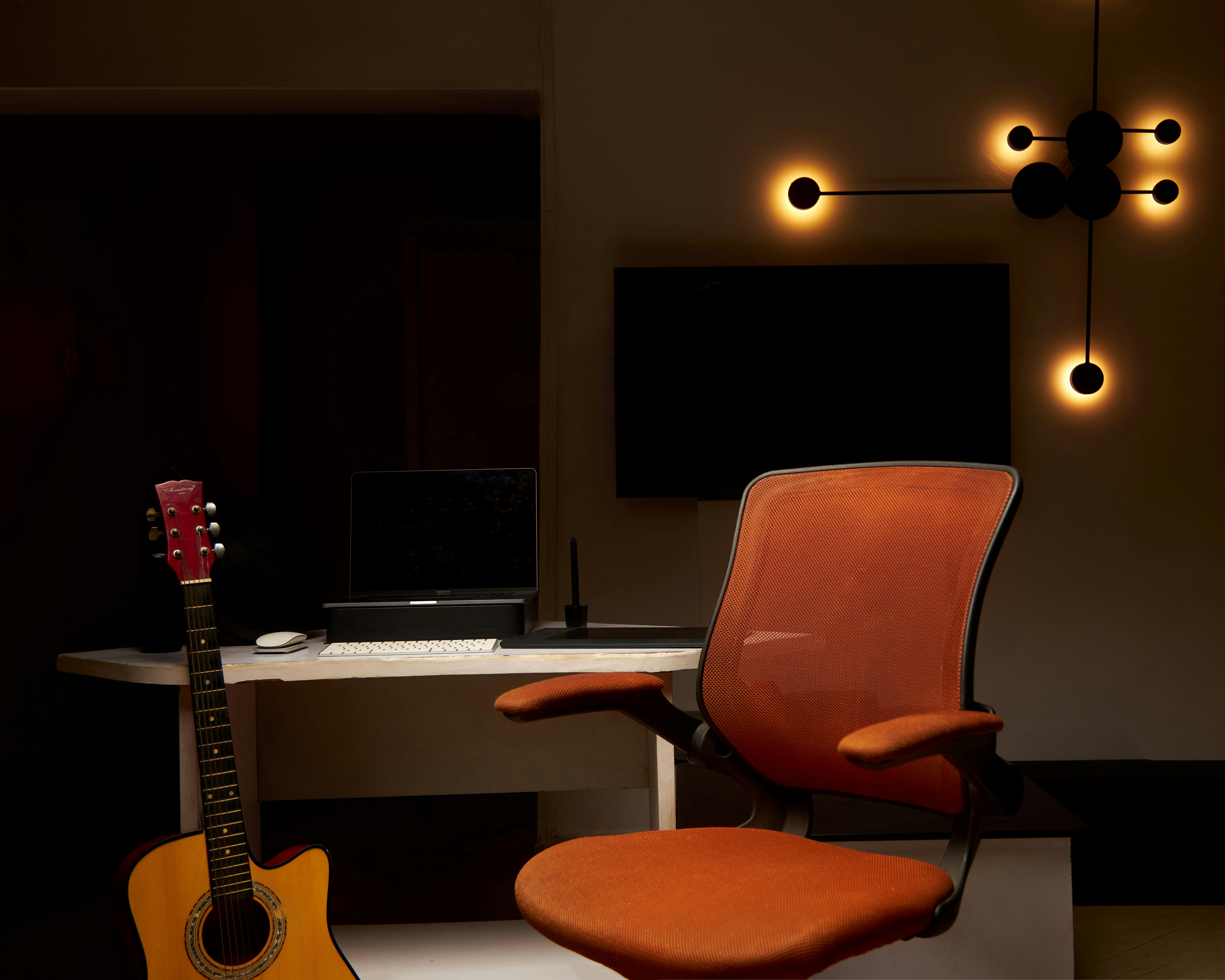 Una silla de oficina de malla naranja junto a un escritorio con una guitarra y un portátil.