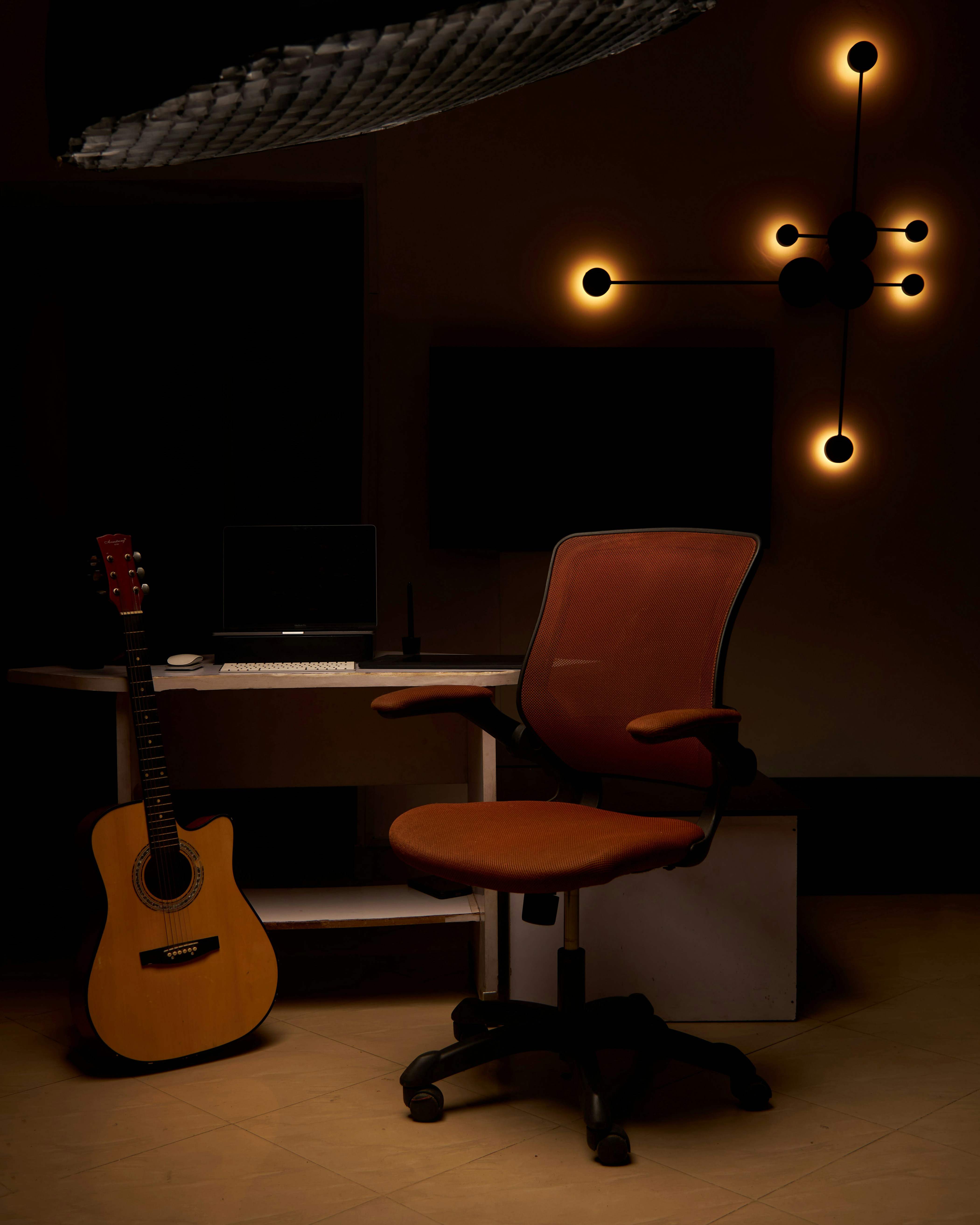 Una guitarra acústica junto a una silla de oficina naranja en un escritorio en un espacio de trabajo.