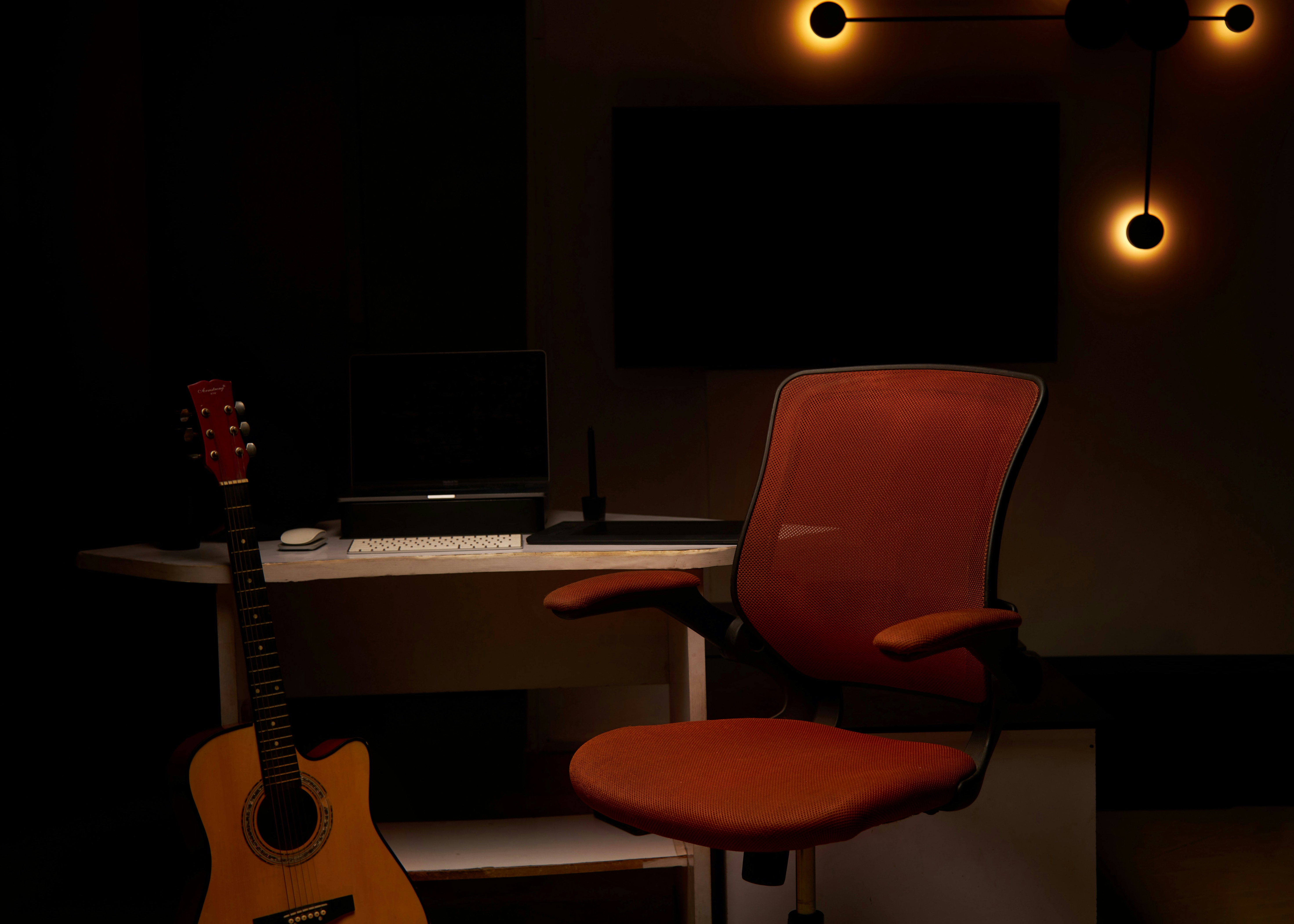 Una guitarra acústica junto a un escritorio con un portátil y una silla naranja de oficina.