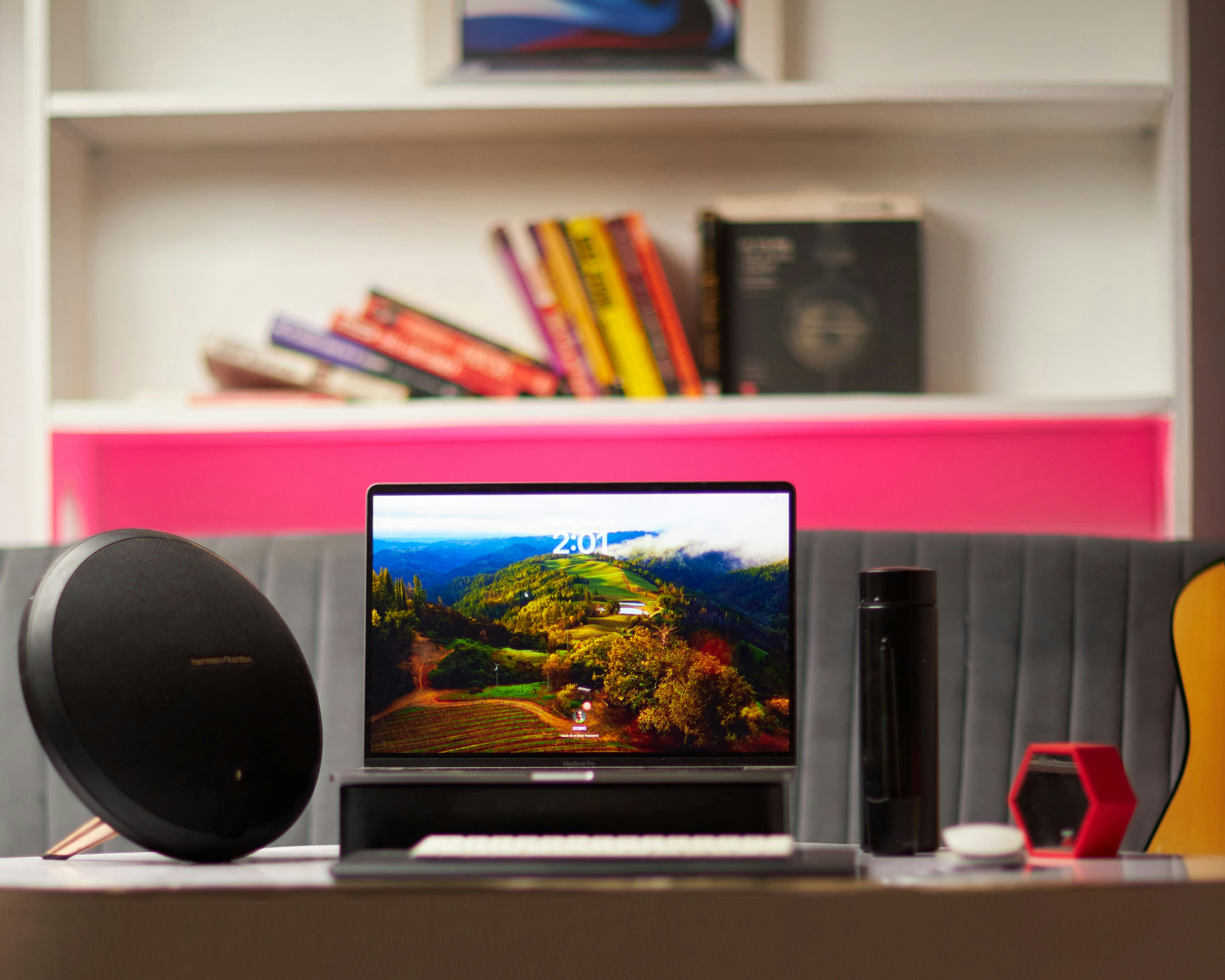 Un portátil sobre un escritorio con altavoz, botella de agua y libros detrás.