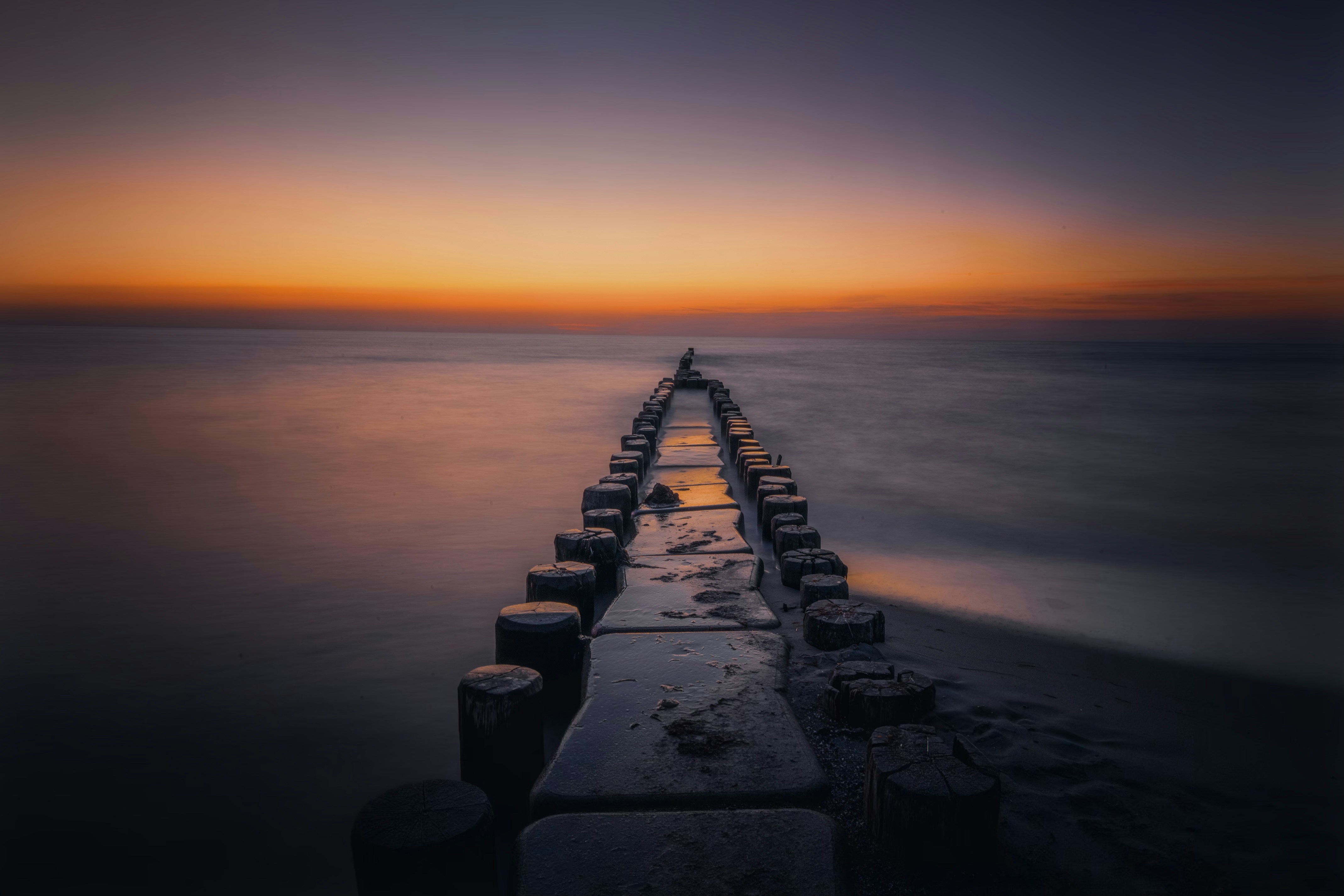 A wooden groyne extending into calm water under a warm orange sky