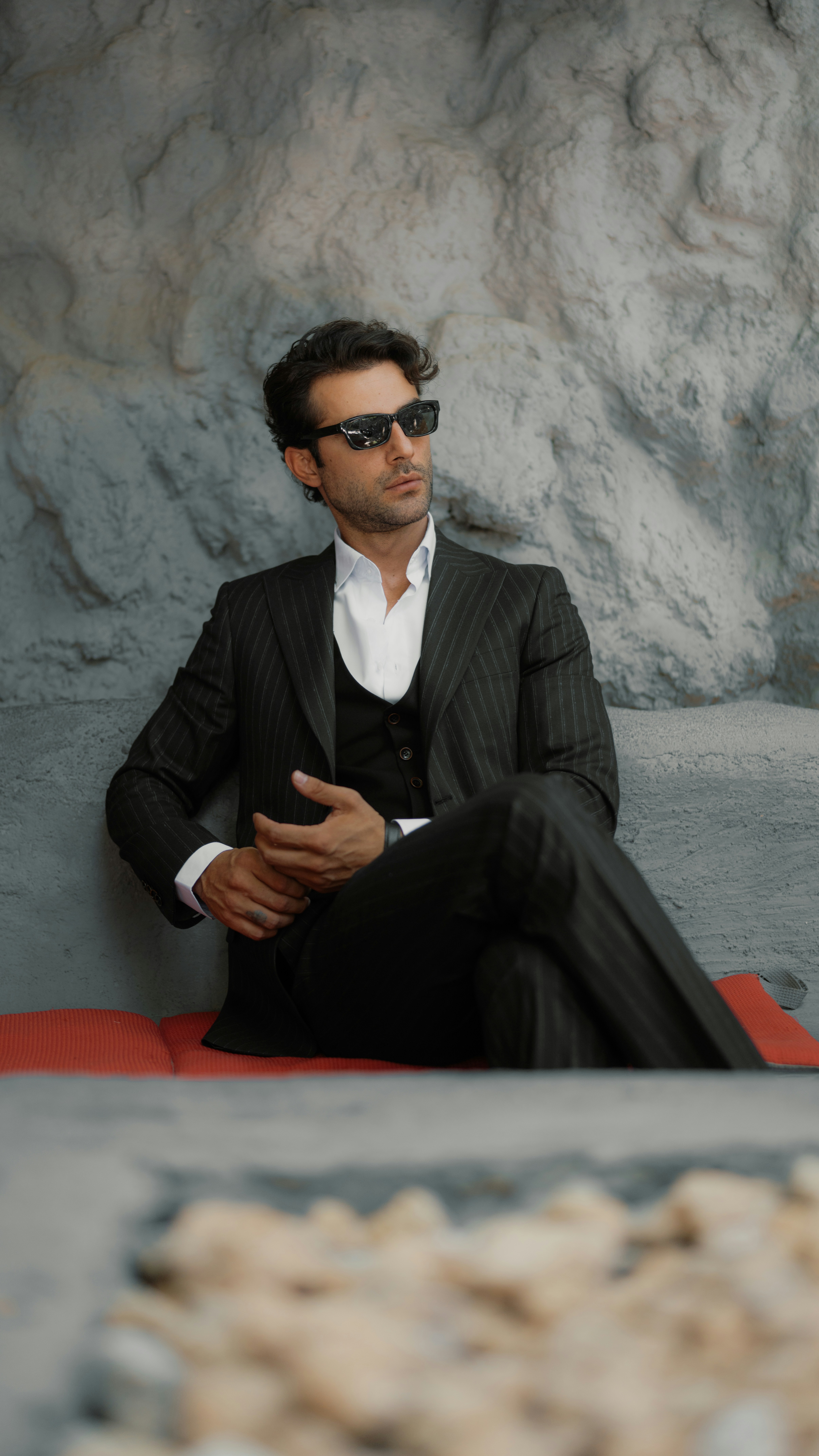 A man in a pinstripe suit and sunglasses against a textured grey stone wall