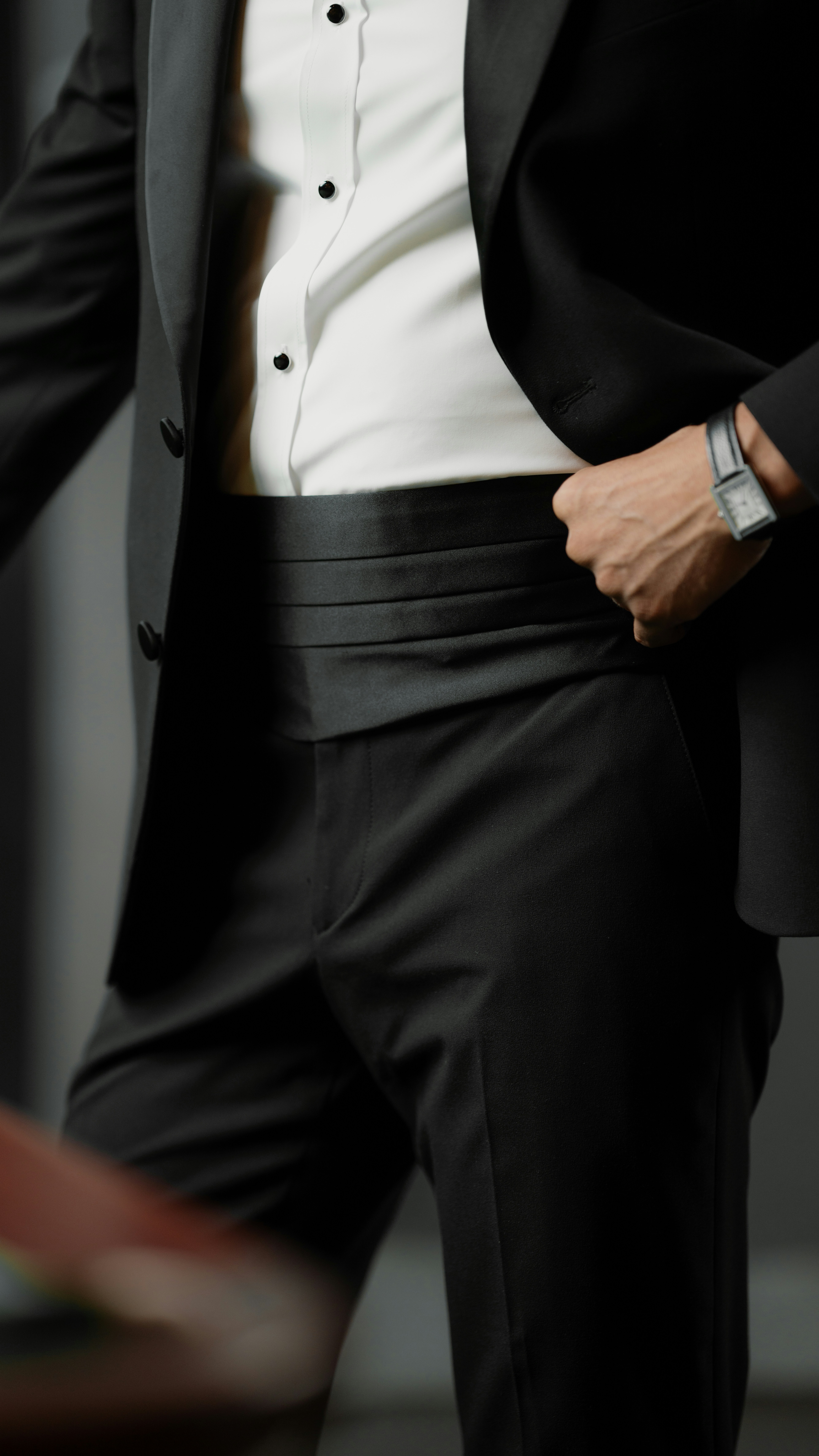 A person in a black tuxedo with a cummerbund and a wristwatch
