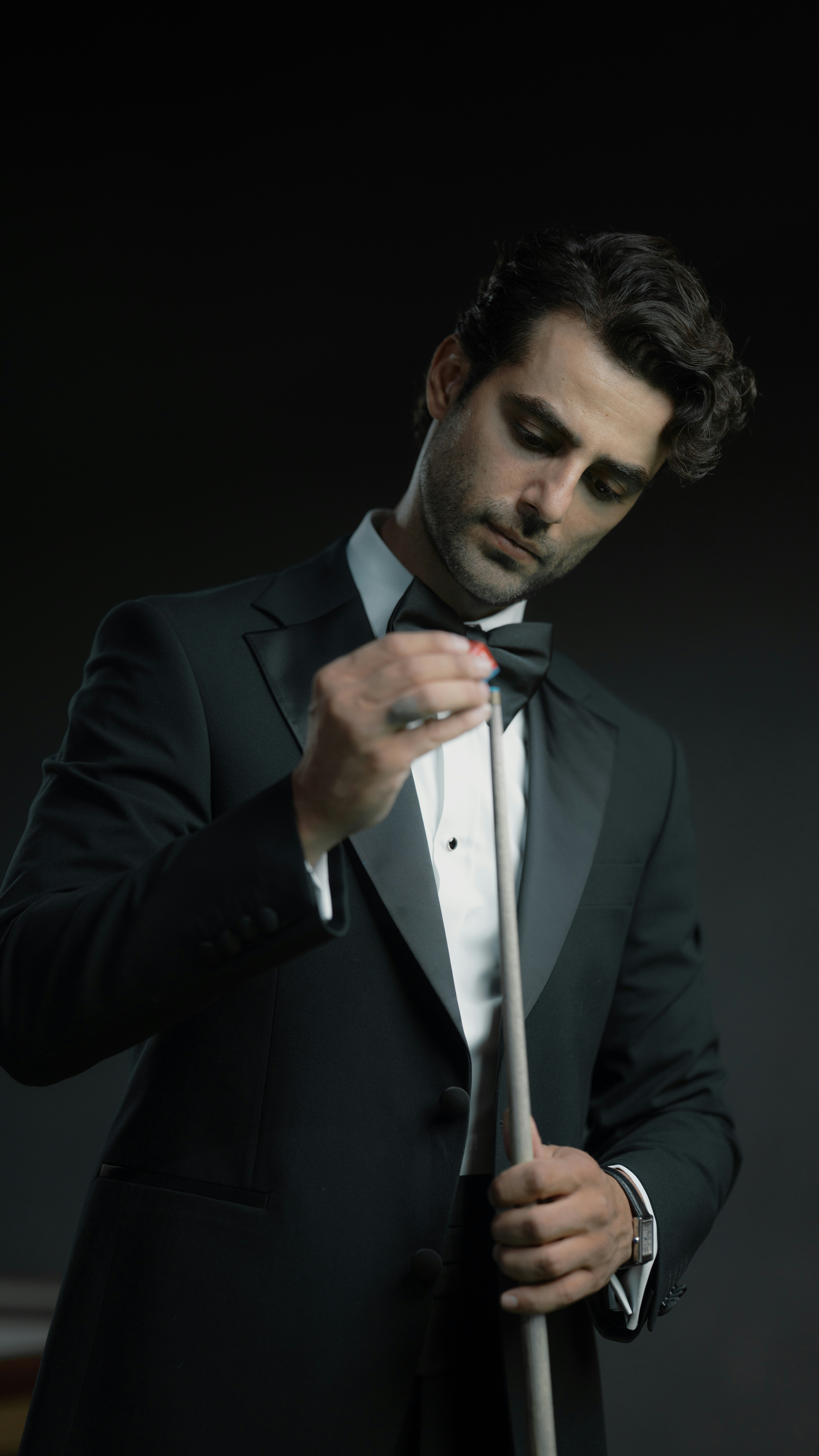 In a tuxedo, chalking a billiard cue