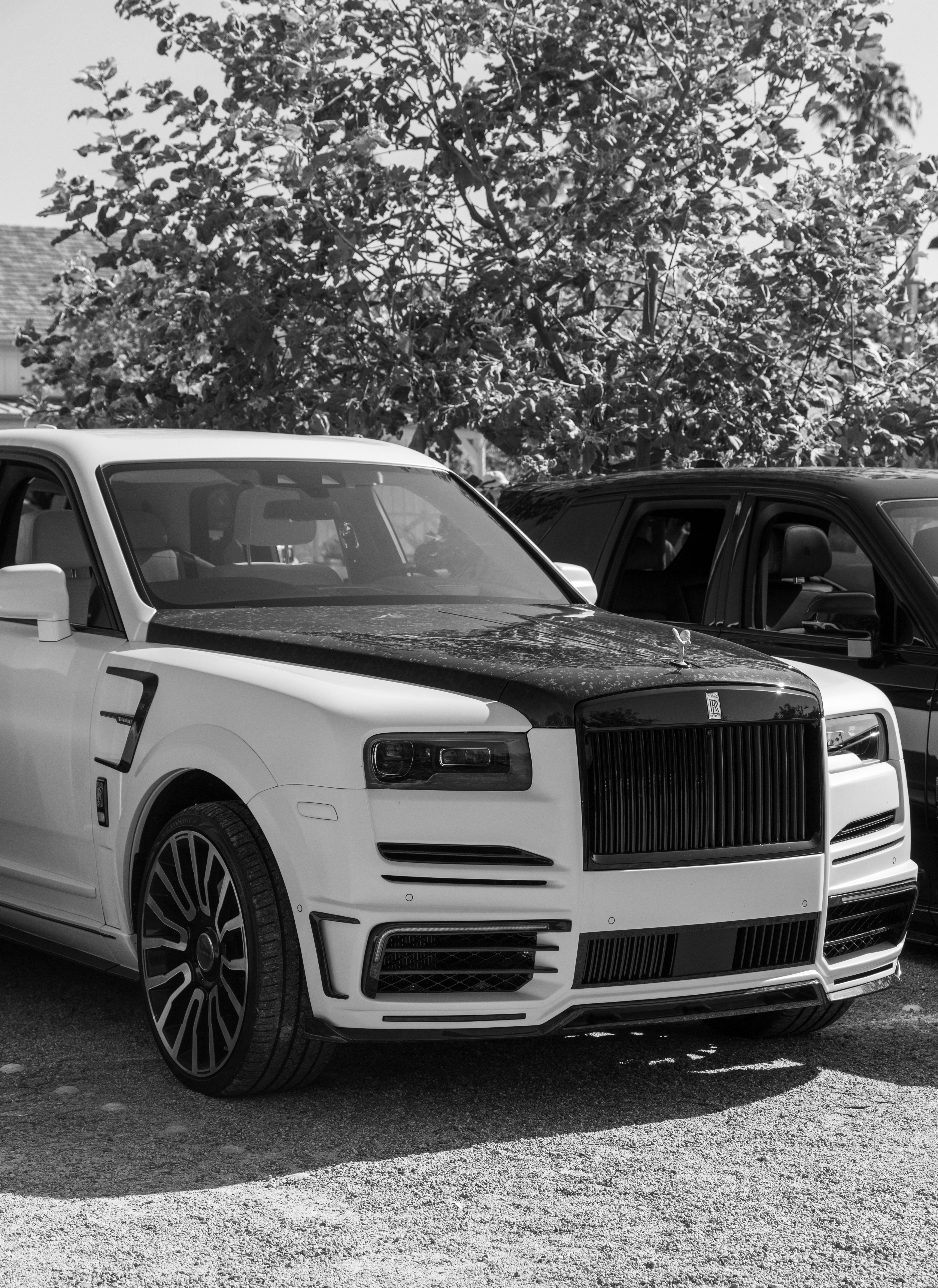 A white Rolls-Royce Cullinan with a black hood parked on a gravel lot.