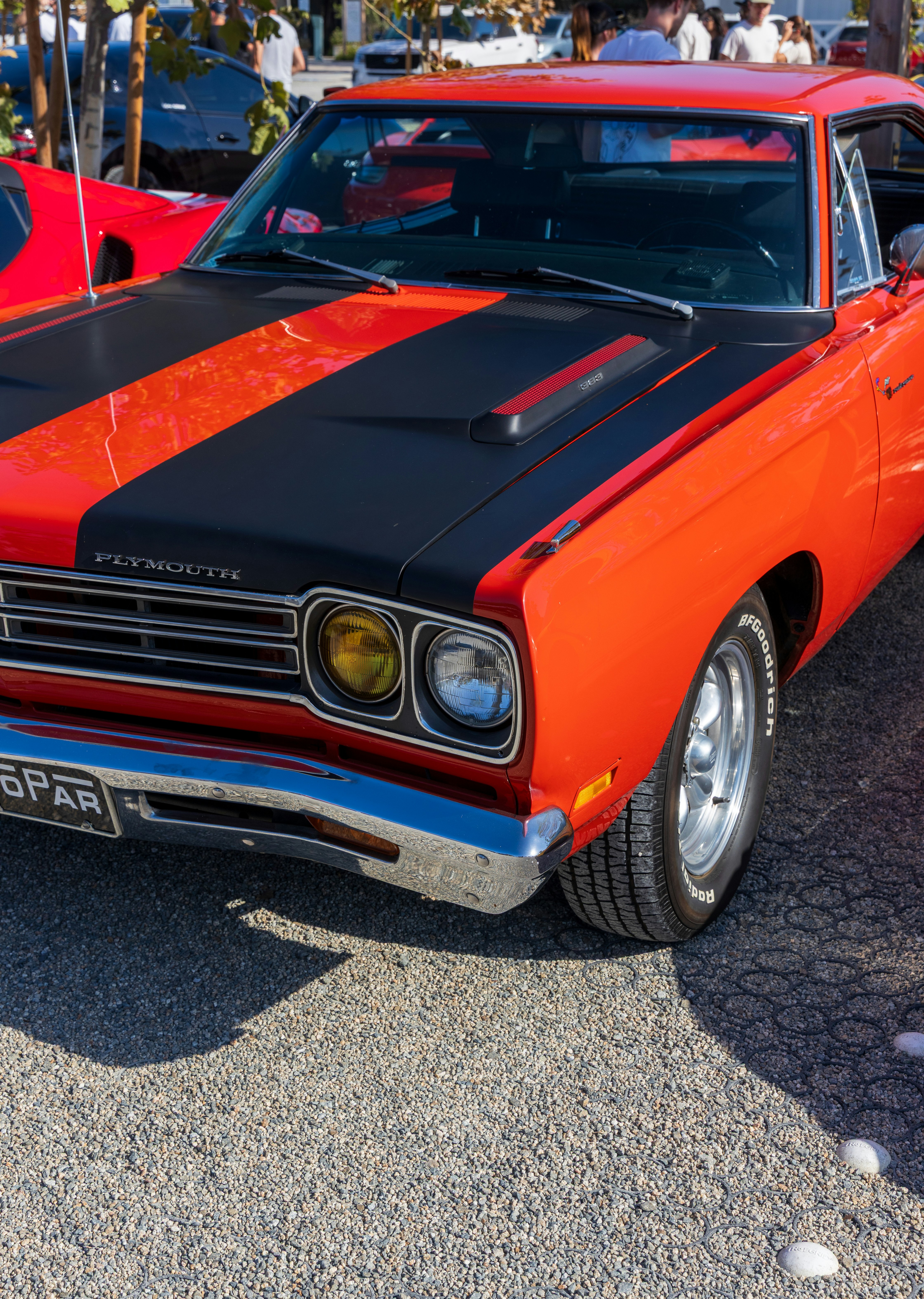 A red Plymouth muscle car with black hood stripes parked on gravel.
