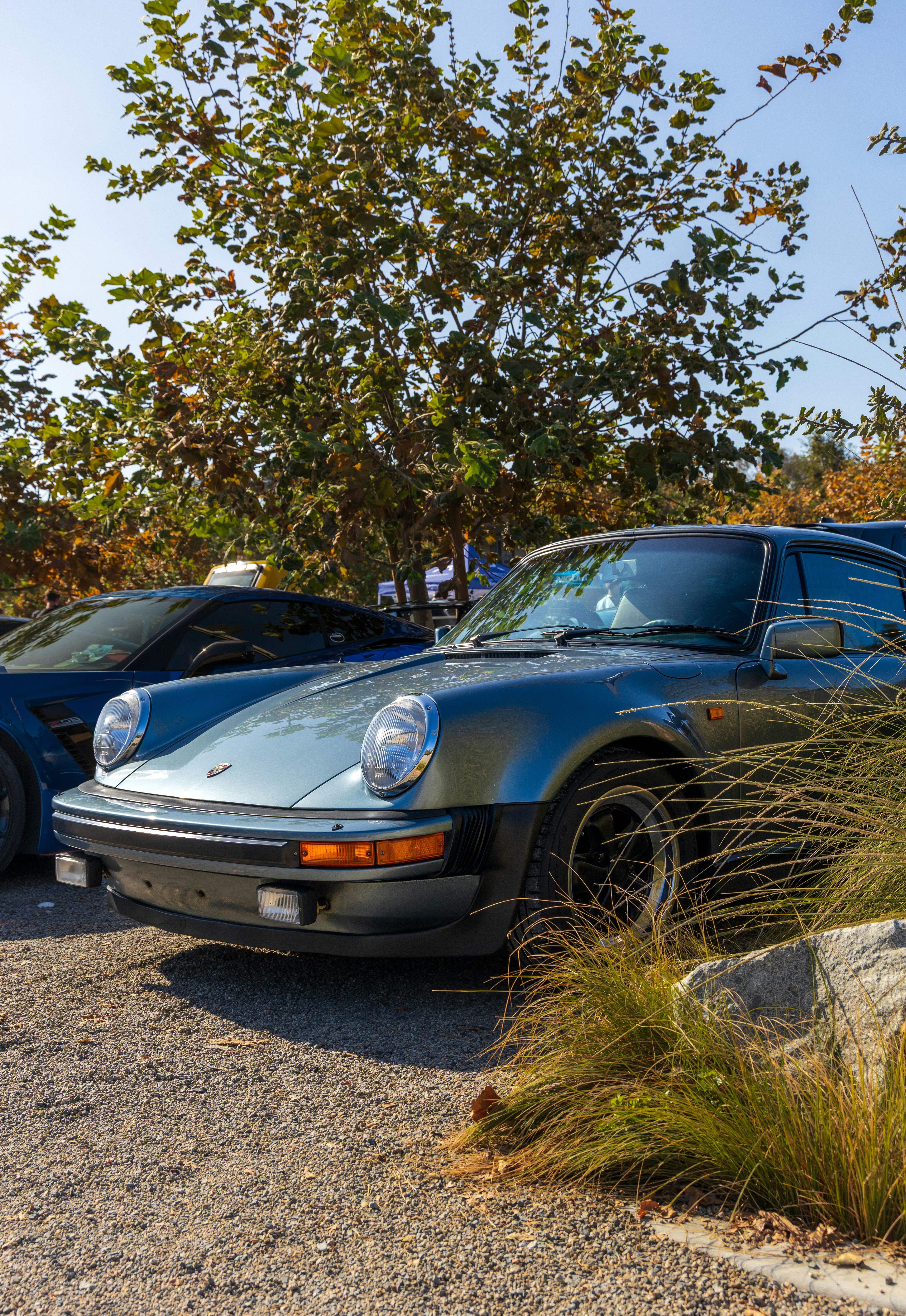 A metallic blue vintage sports car parked on a gravel lot near trees.