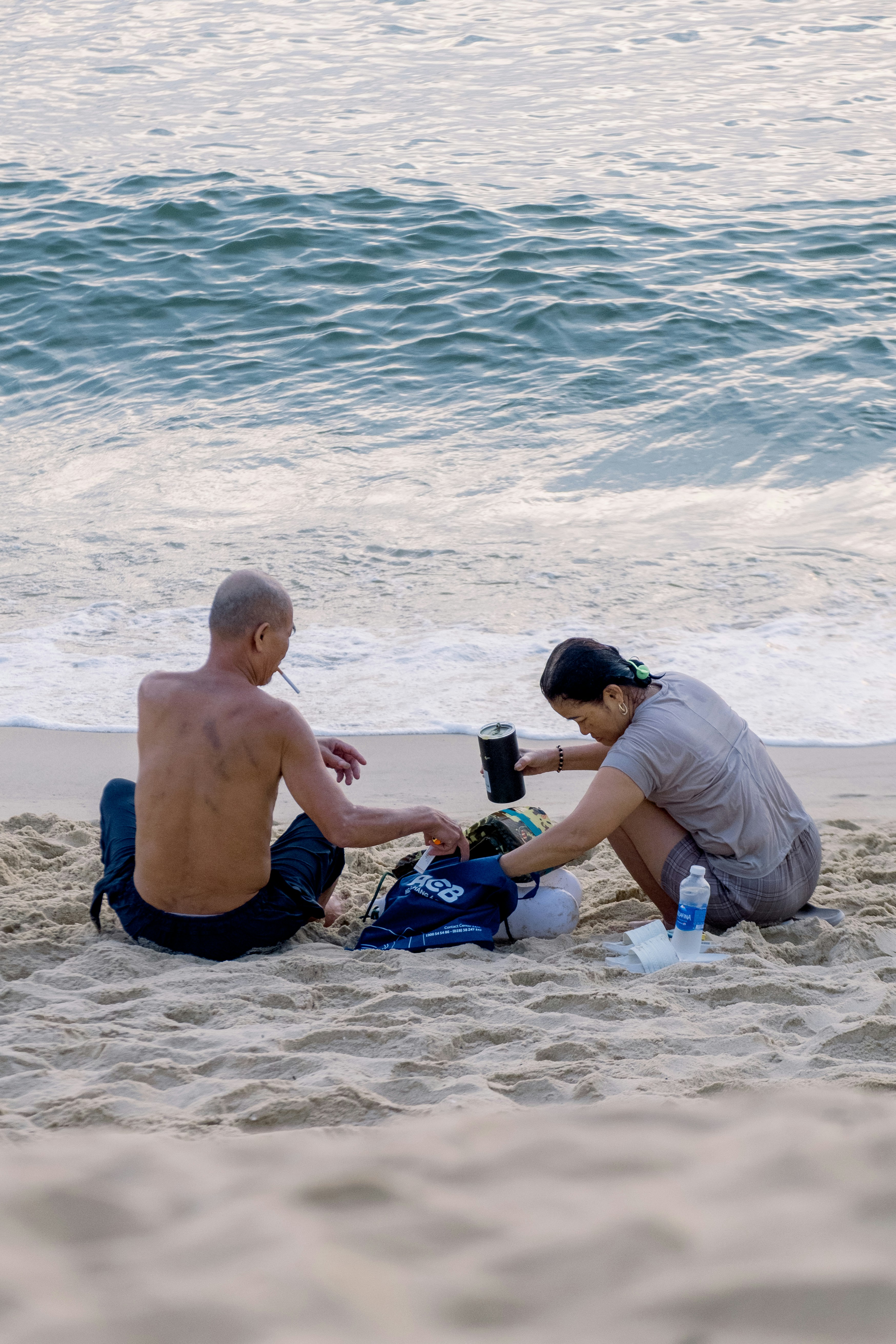 A man and a woman sitting on a sandy beach by the ocean.