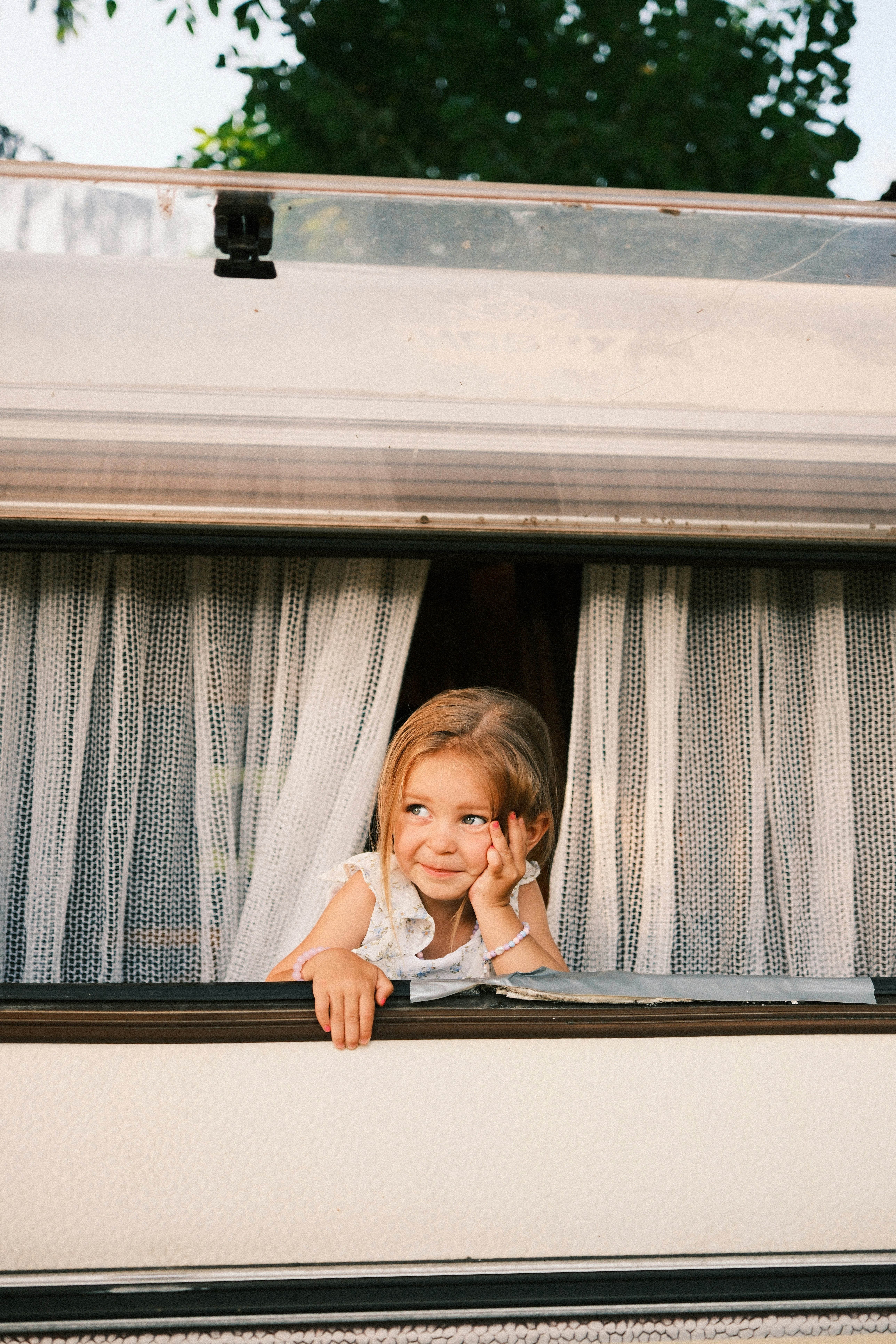A young child leaning out of a caravan window with white curtains.