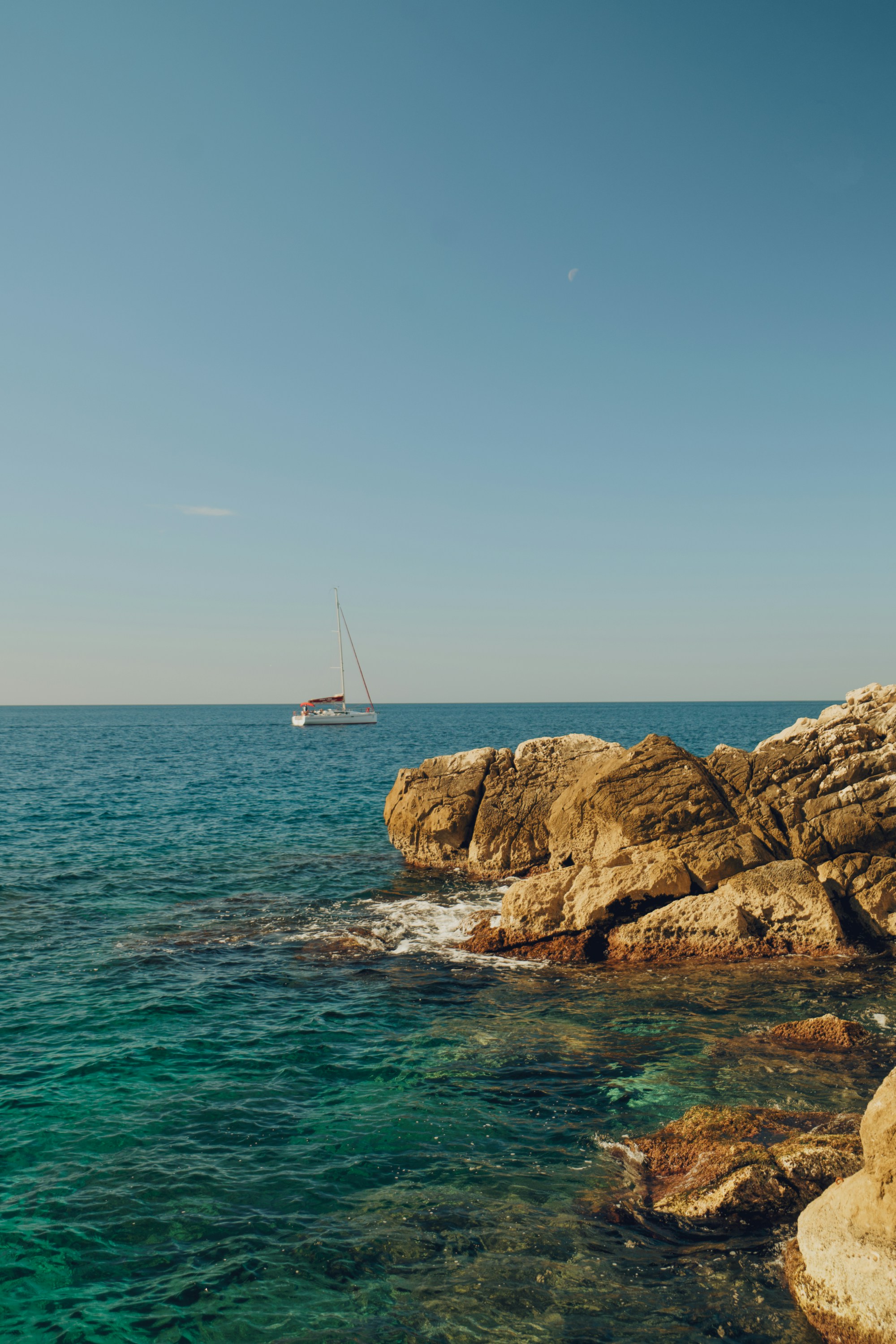 A sailboat on the blue sea near a rocky coastline under a clear sky.
