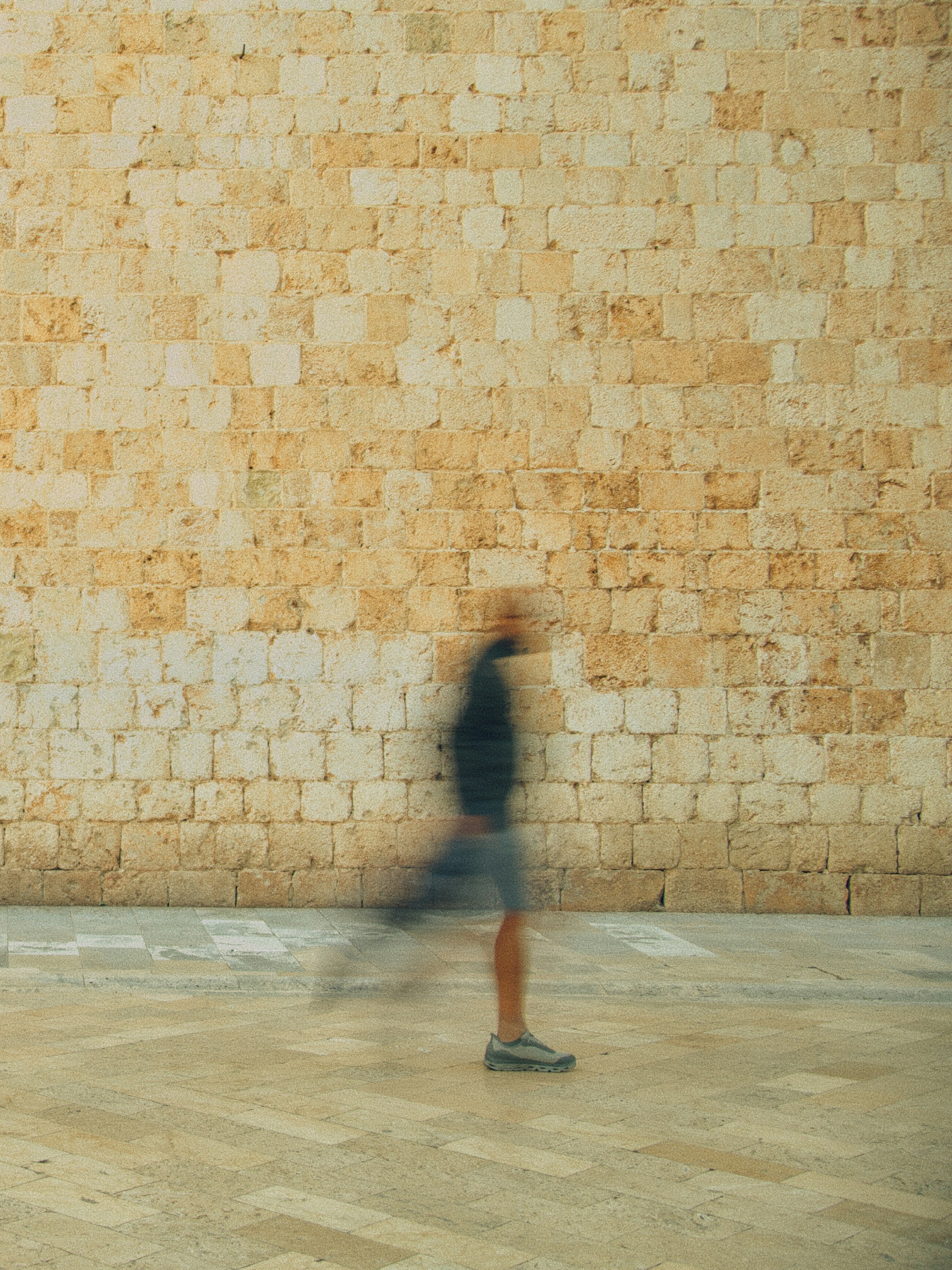 A blurred person walking past a textured stone wall on a paved plaza.