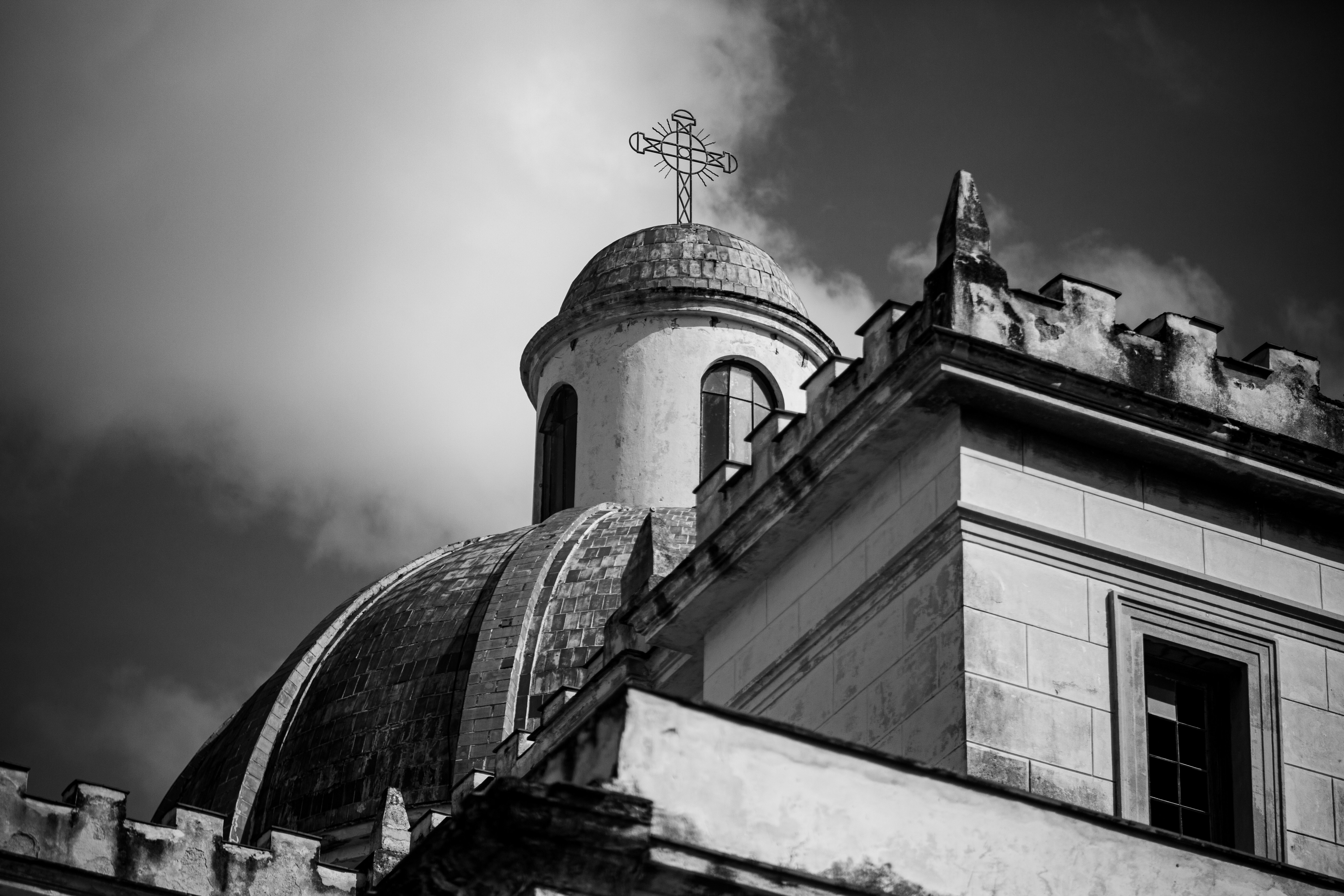 A metal cross atop a domed church roof against a cloudy sky.