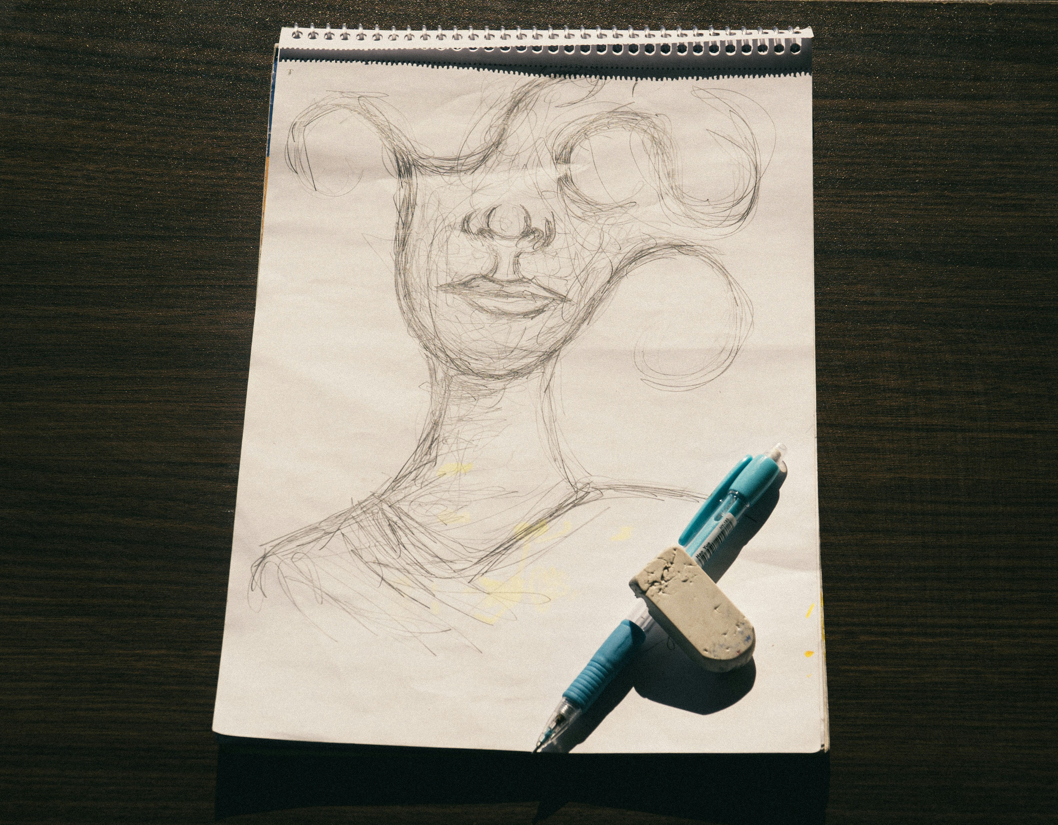 A pencil sketch of a face on a notepad with a blue pen and eraser.