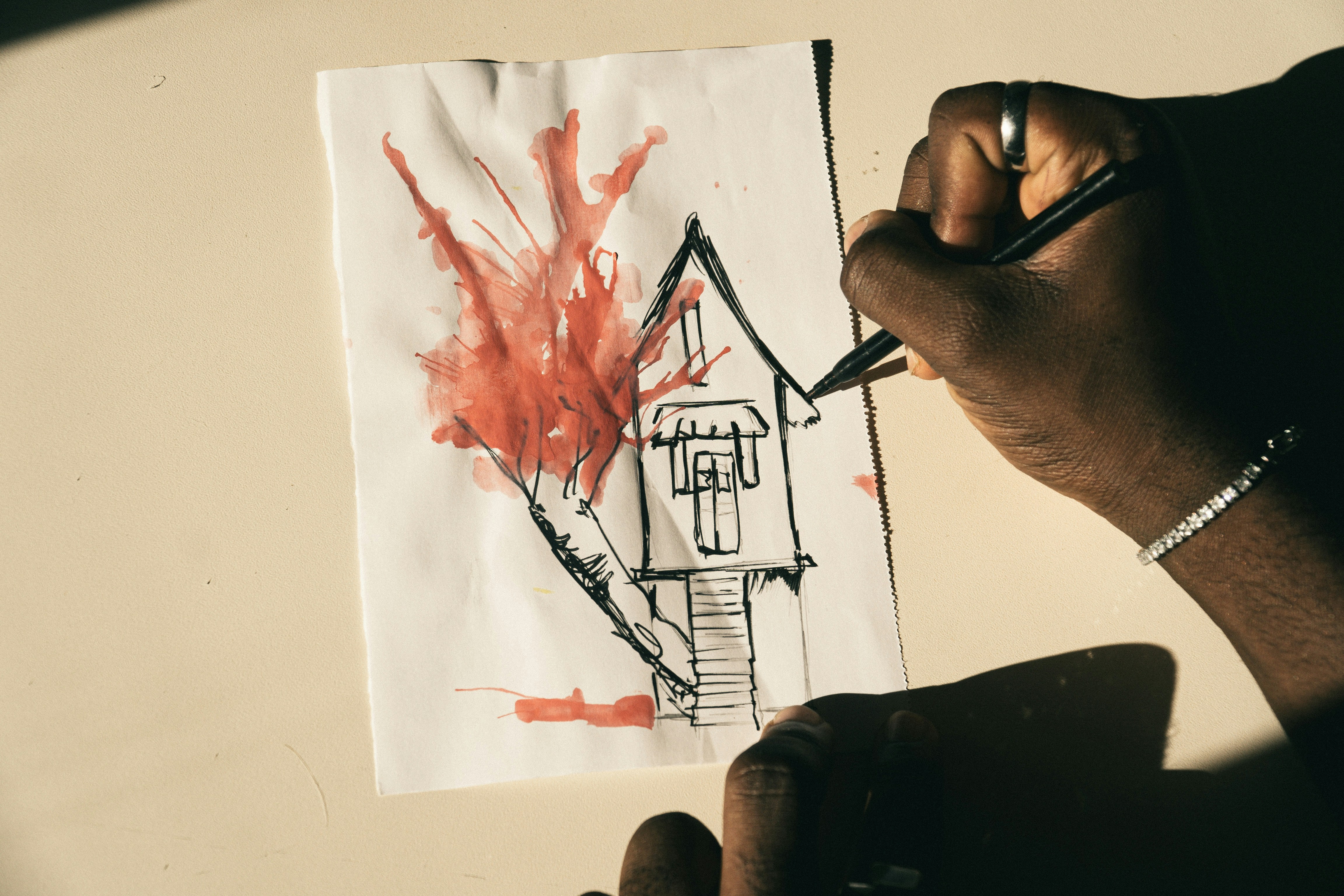 A person drawing a house with a red watercolor tree on white paper.