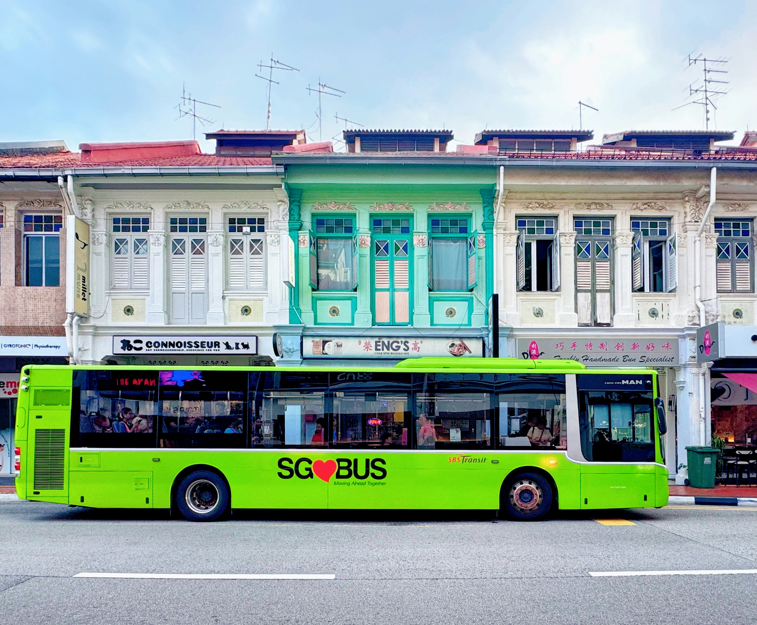 A bright green public bus driving past colorful historic shophouses in Singapore.
