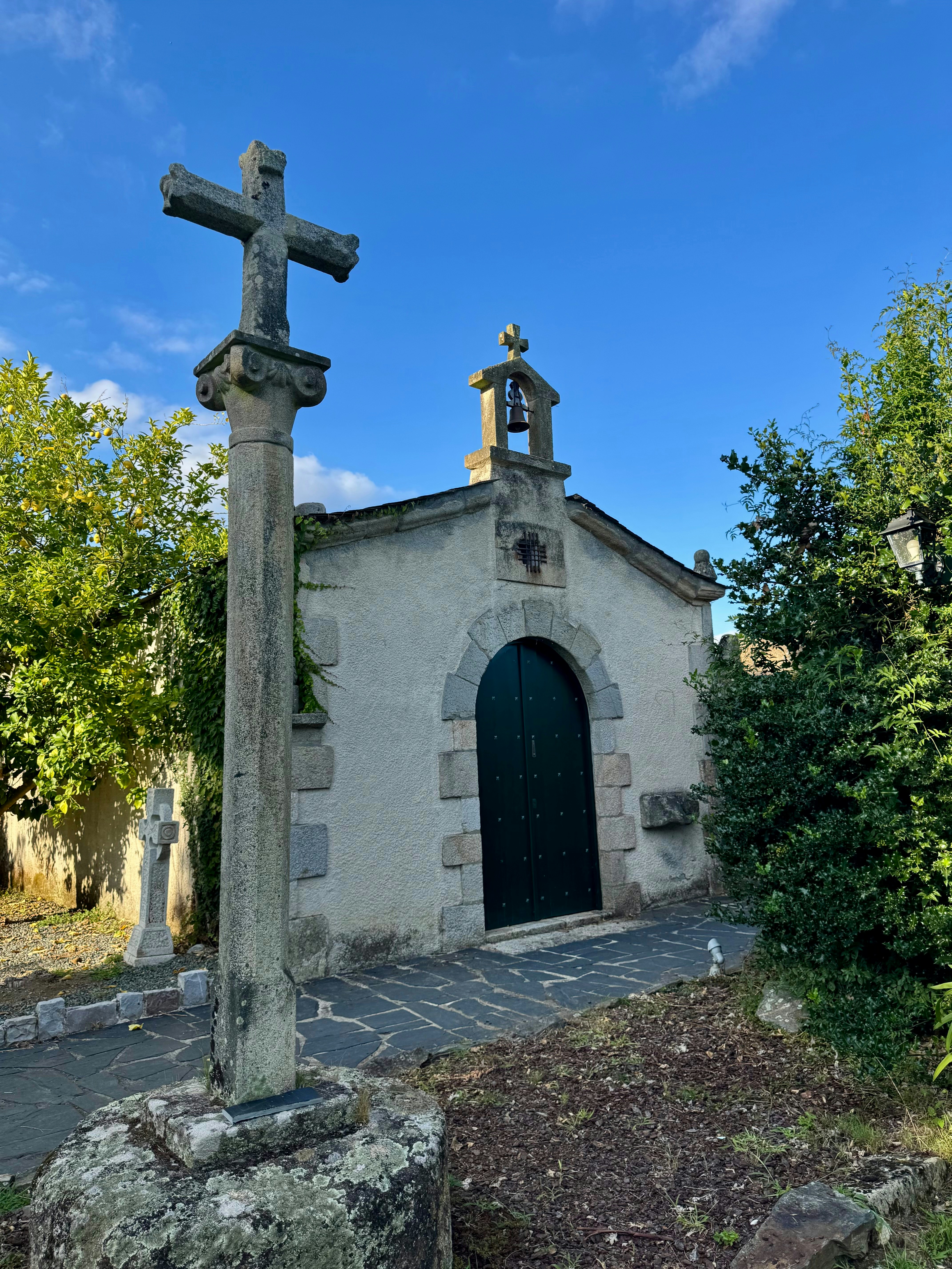 A stone cross on a pillar before a small chapel with a bell tower.