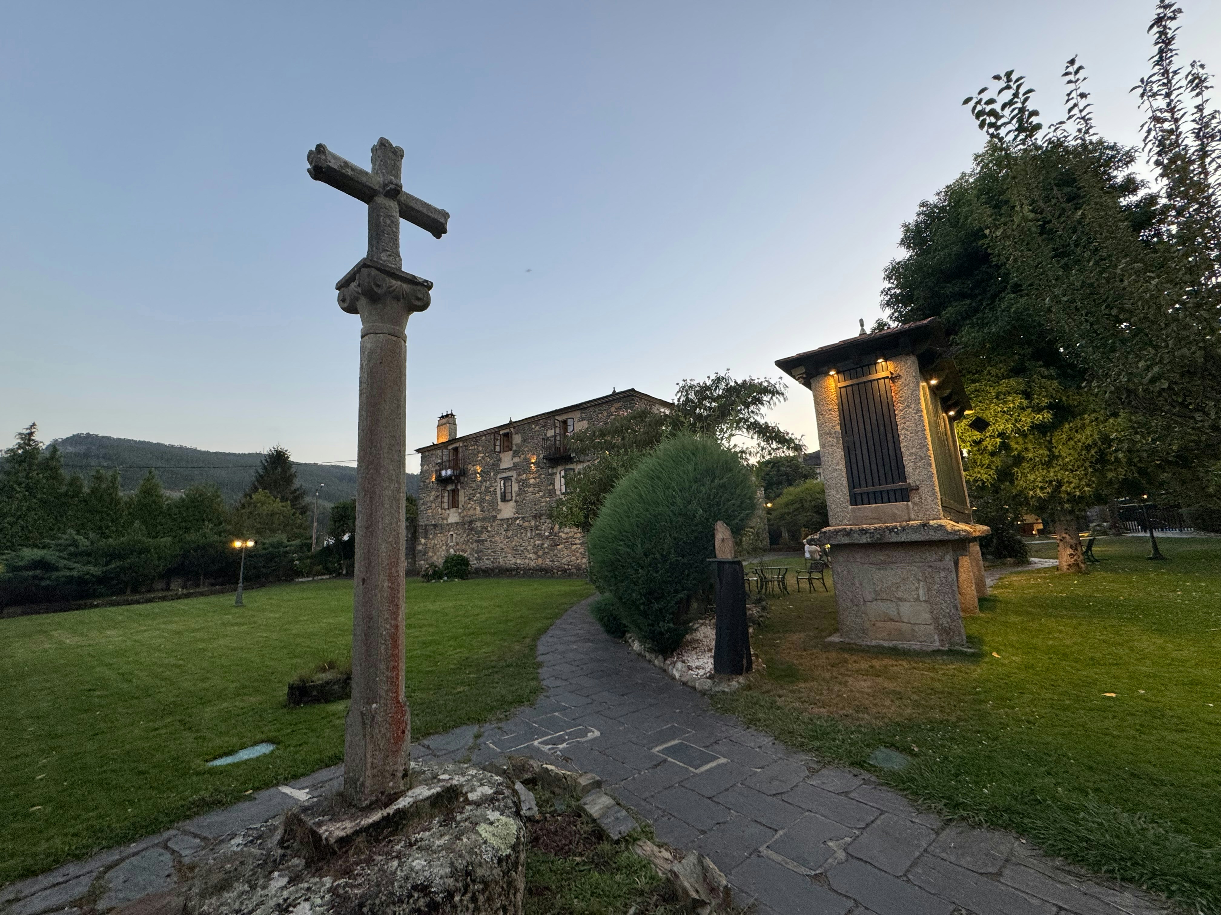 A stone cross on a pedestal in a garden with a stone granary.