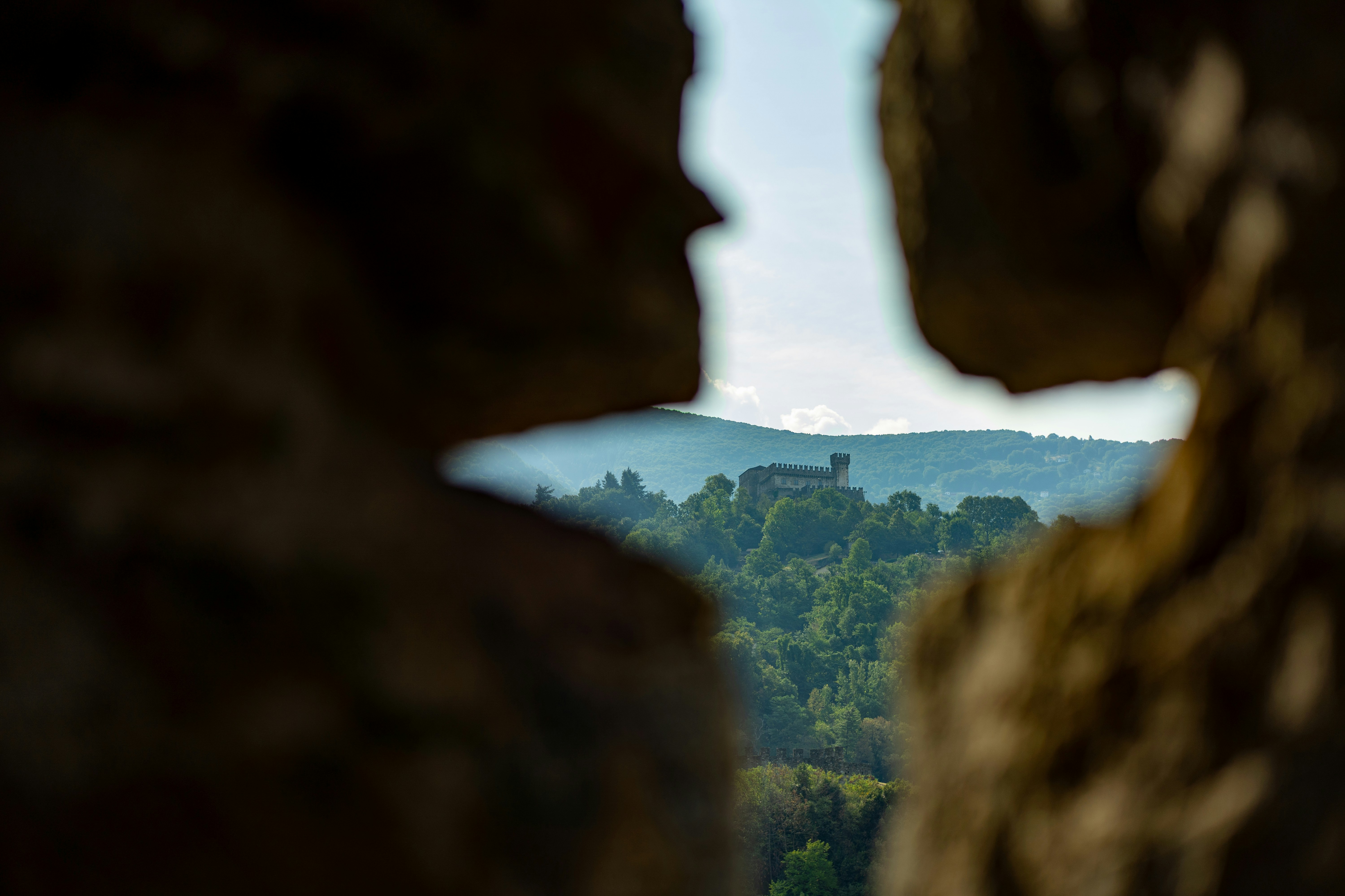 Um castelo de pedra em uma colina arborizada visto por uma estreita abertura de pedra.
