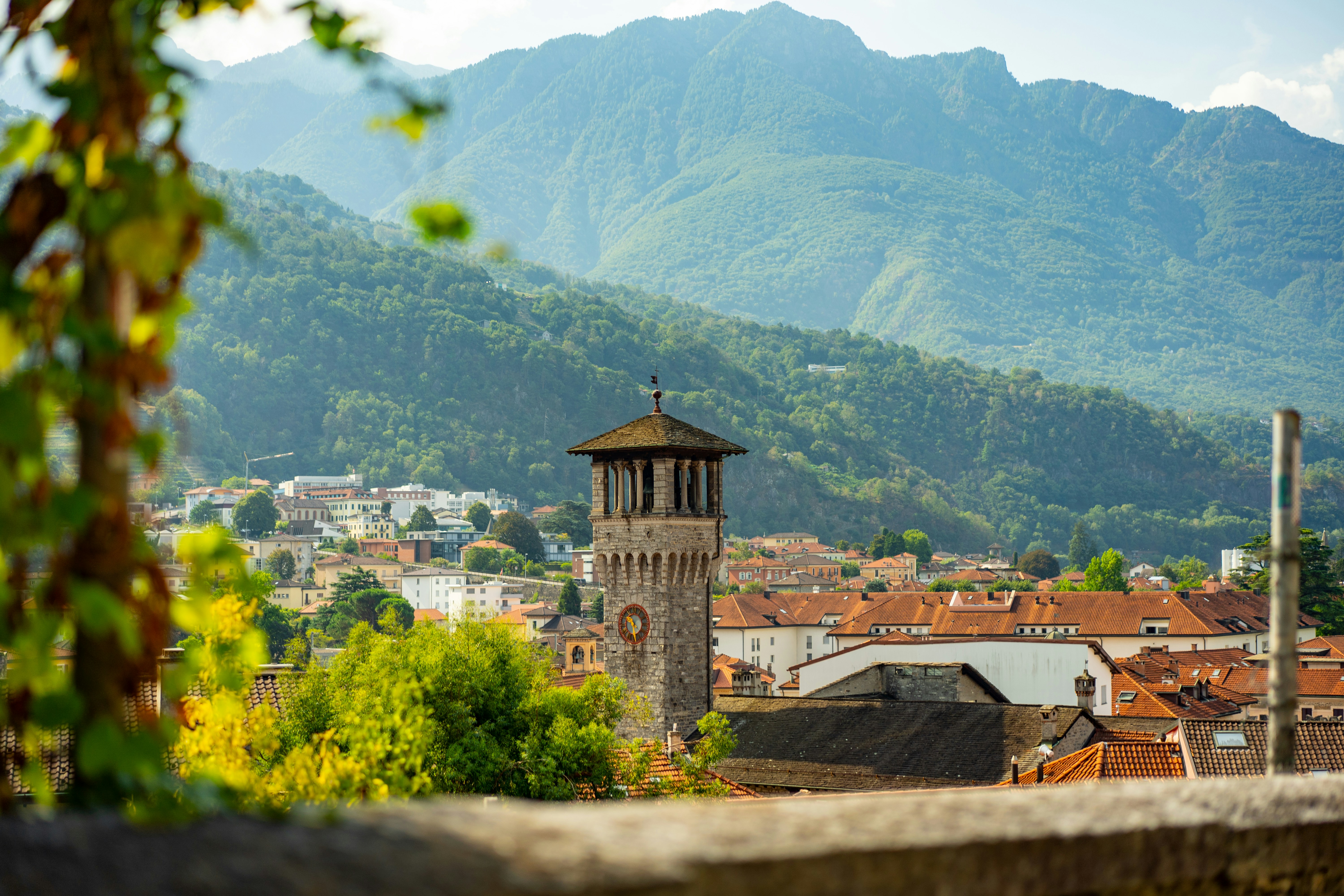 Uma torre de relógio de pedra se erguia acima de uma cidade com montanhas ao fundo.