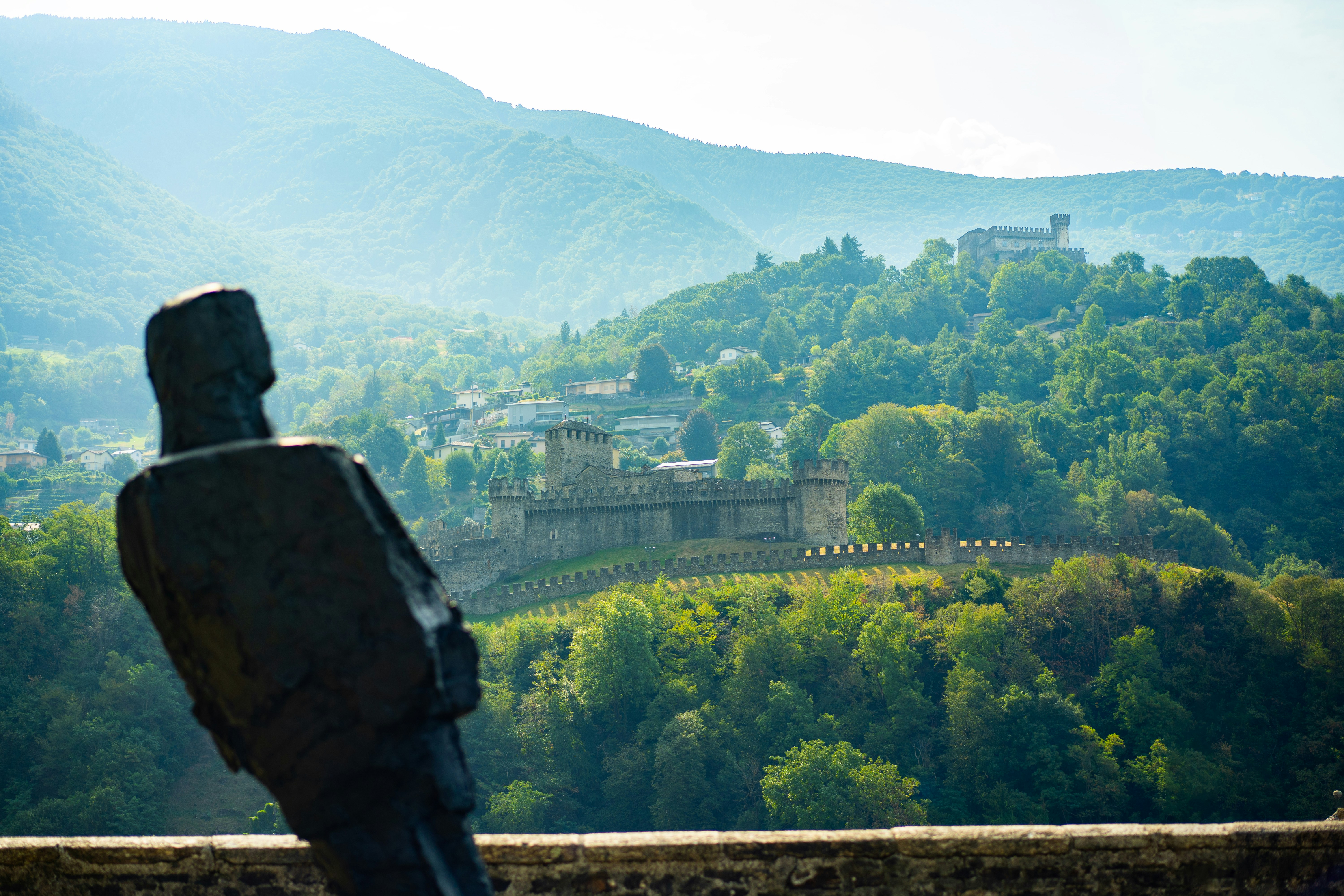 Uma escultura de metal escuro com vista para a fortaleza medieval de Castelgrande em uma encosta verde.