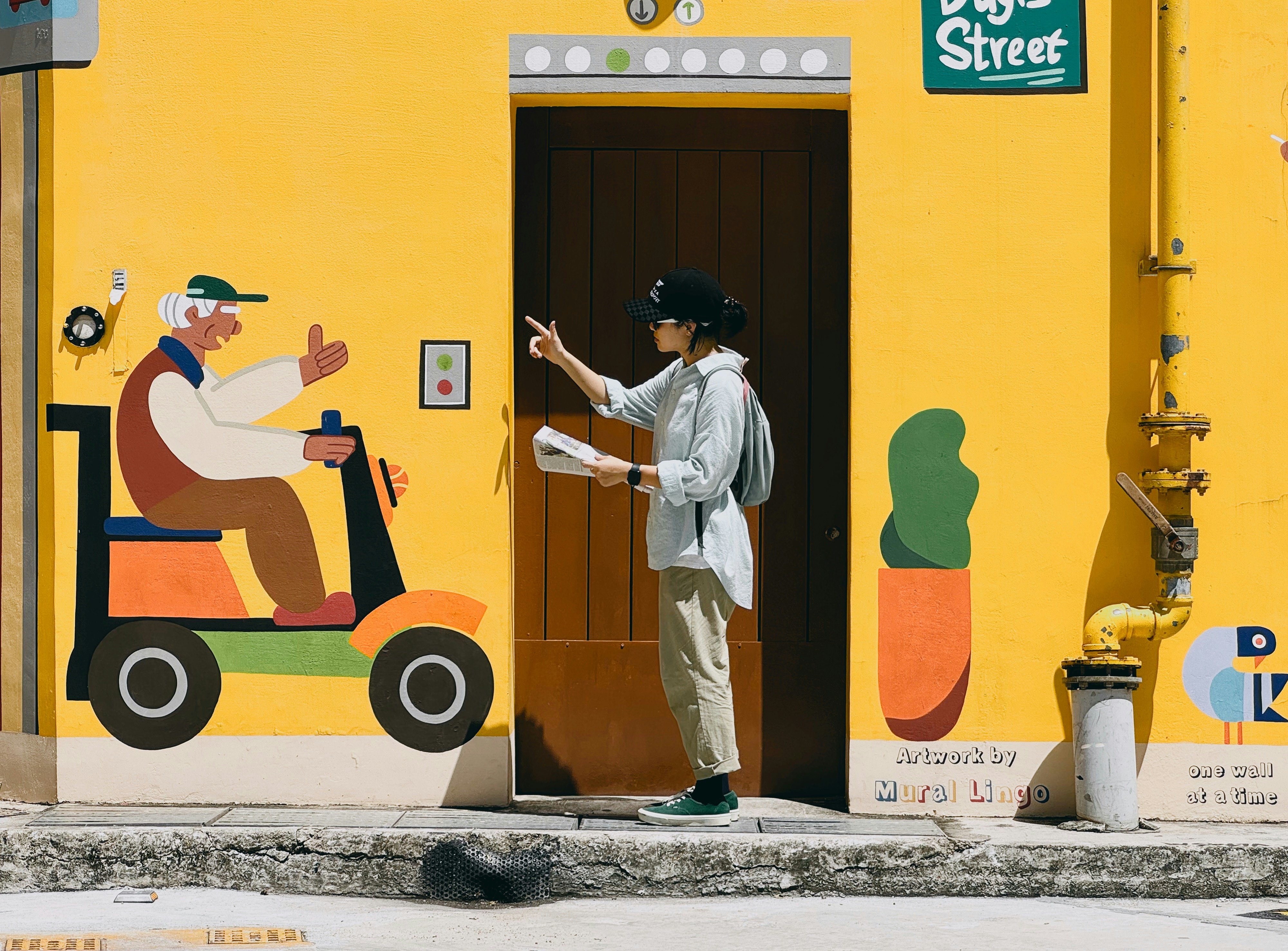 A person in a cap and light shirt pointing at a yellow mural.