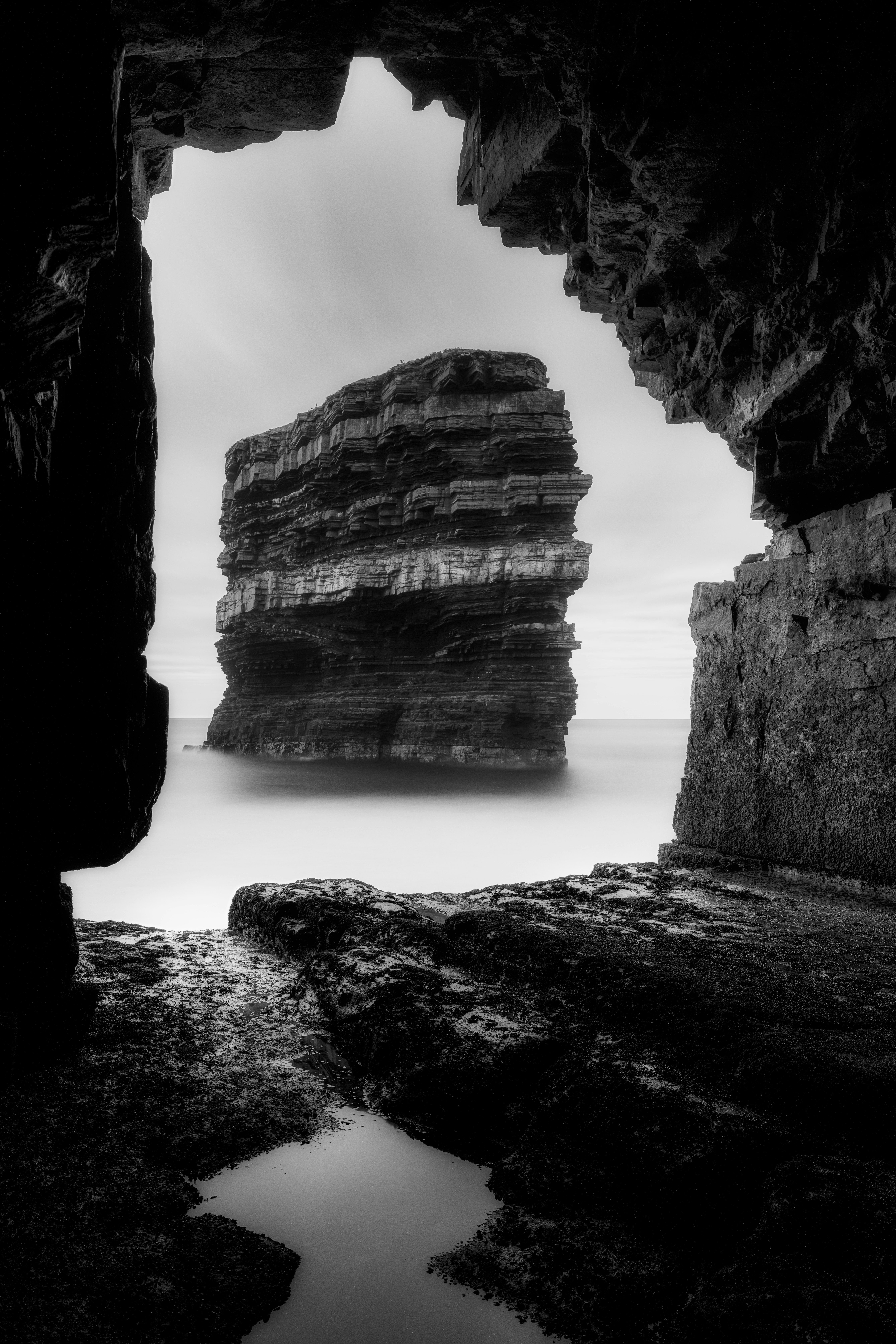 A tall sea stack viewed through a dark rocky cave opening