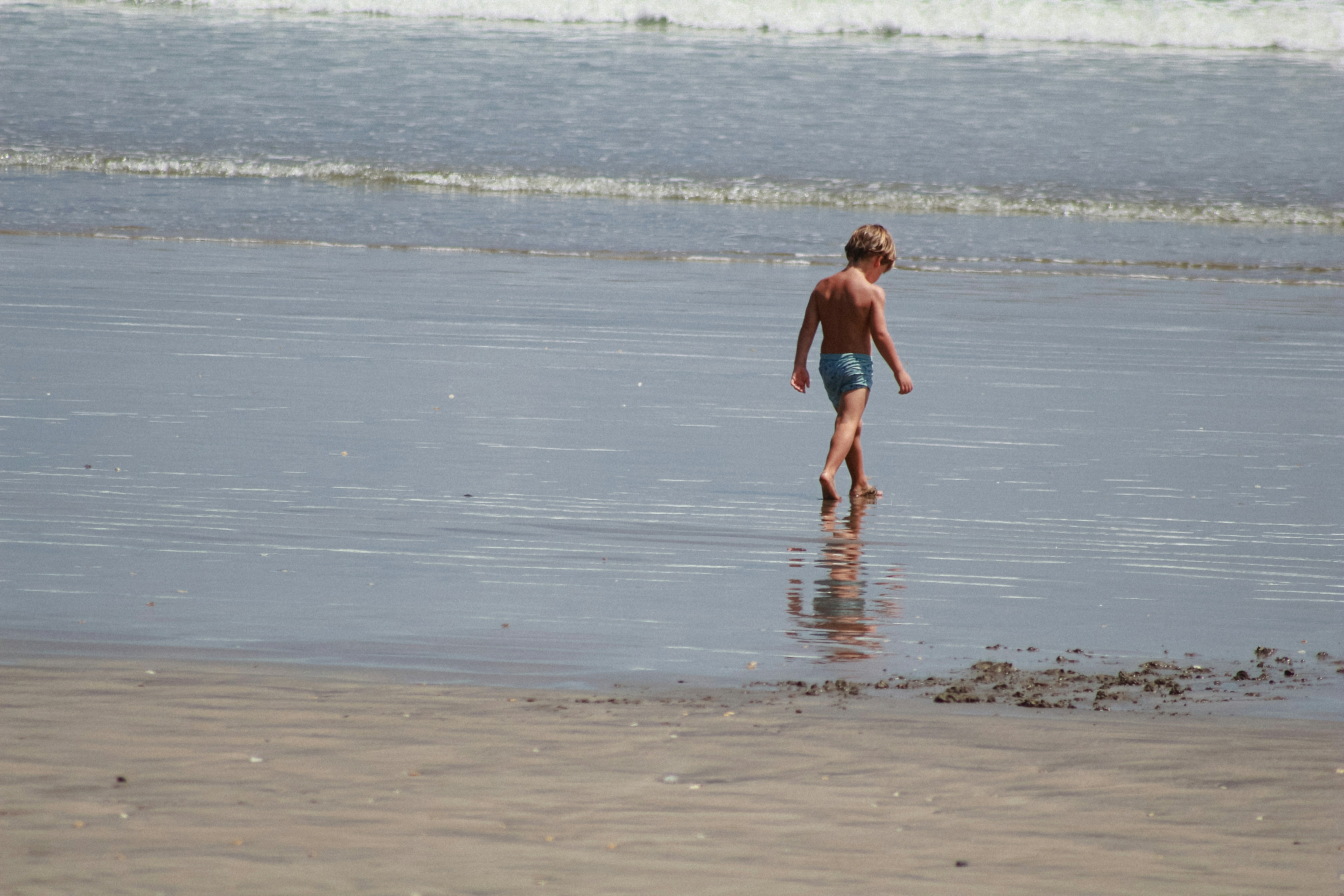 Ein Kind in blauen Badehosen, das durch flaches Wasser an einem Sandstrand geht