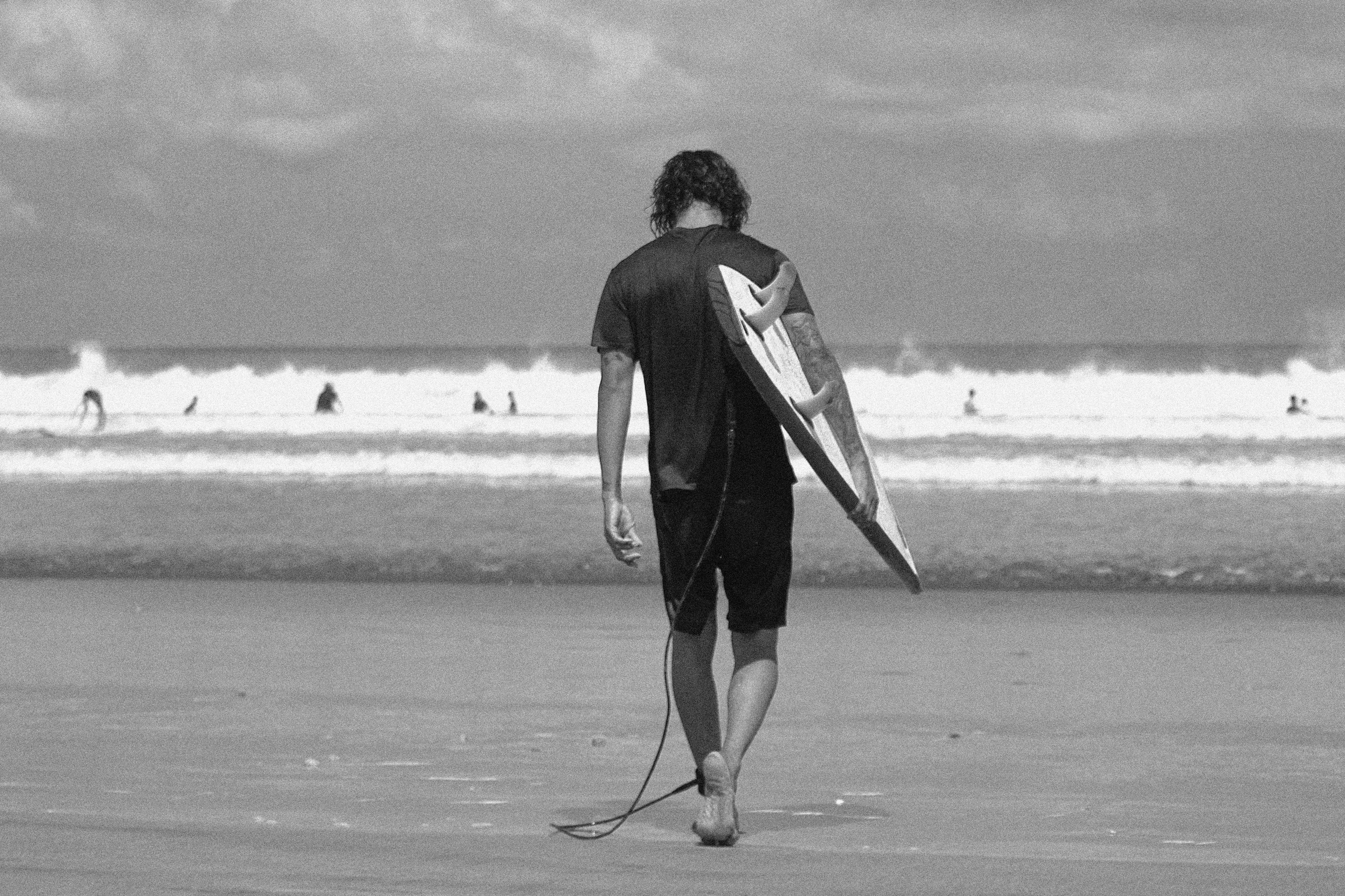 Eine Person mit langen Haaren, die ein Surfbrett an einem Sandstrand trägt