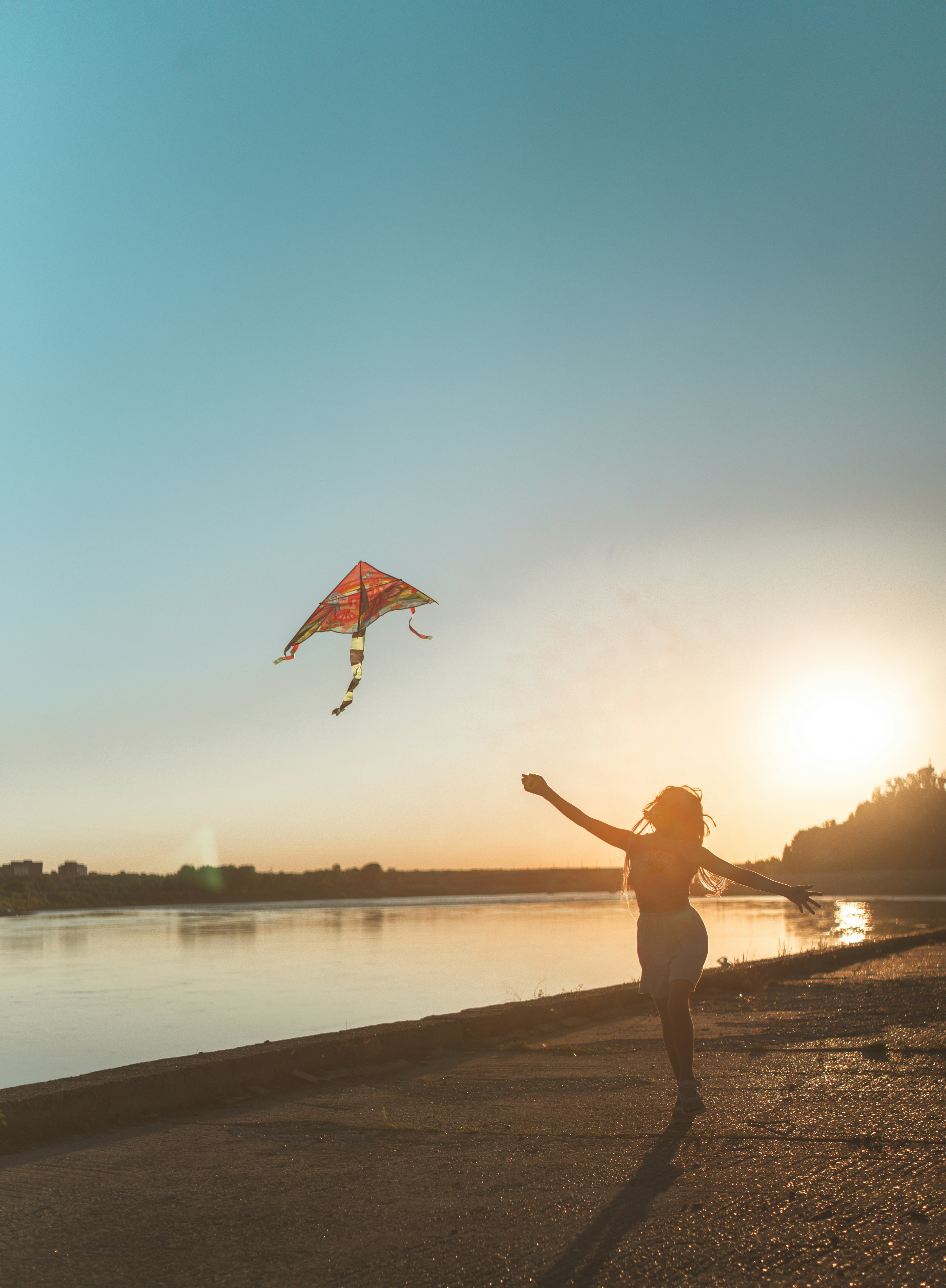 Une personne en silhouette courant avec un cerf-volant coloré près d’une rivière au bas du soleil.