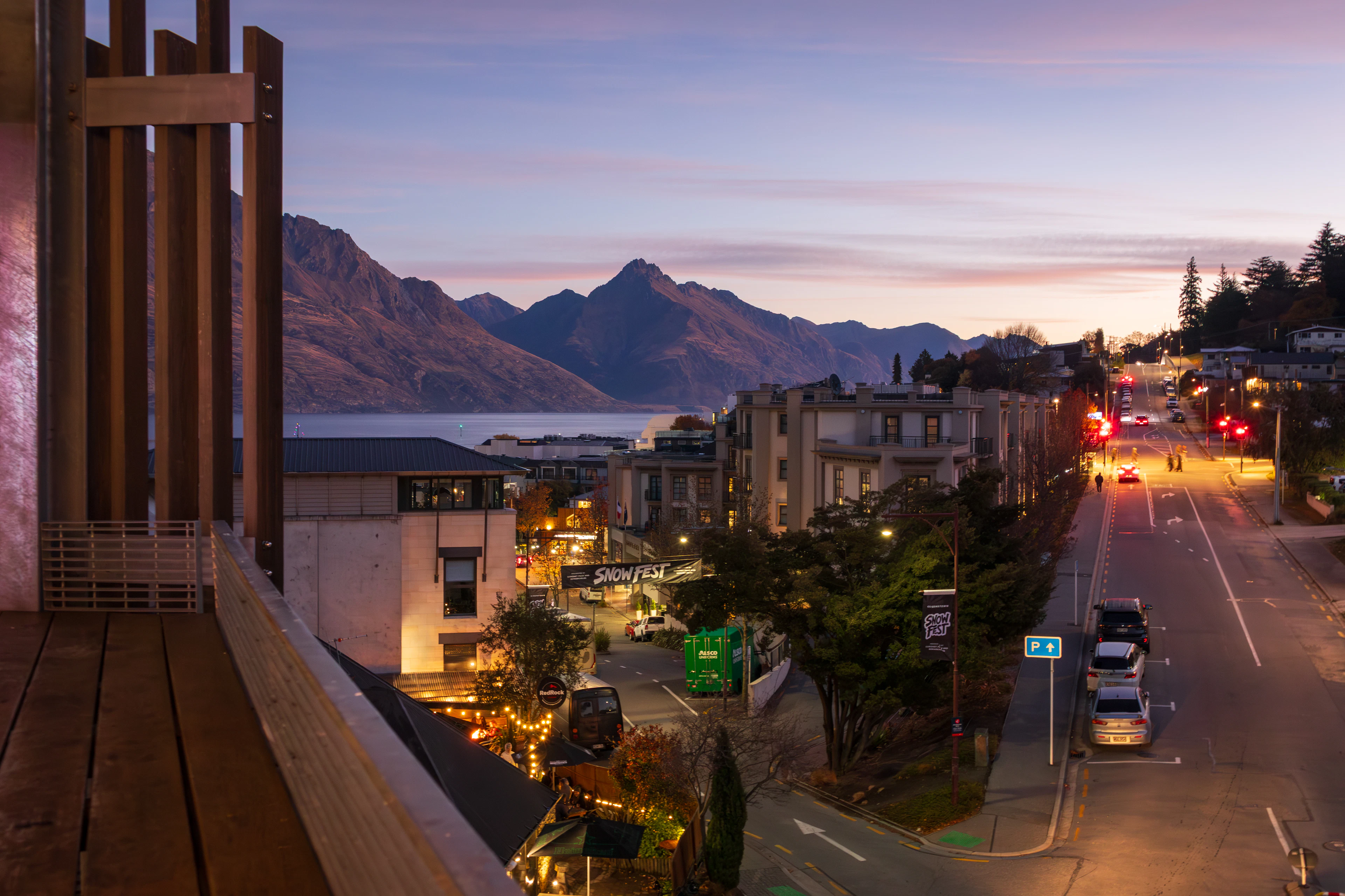 Queenstown's Shotover Street strip glowing at dusk seen from a balcony above, lit shopfronts and a quiet road below, with the dark peaks across Lake Wakatipu holding the last pink light.