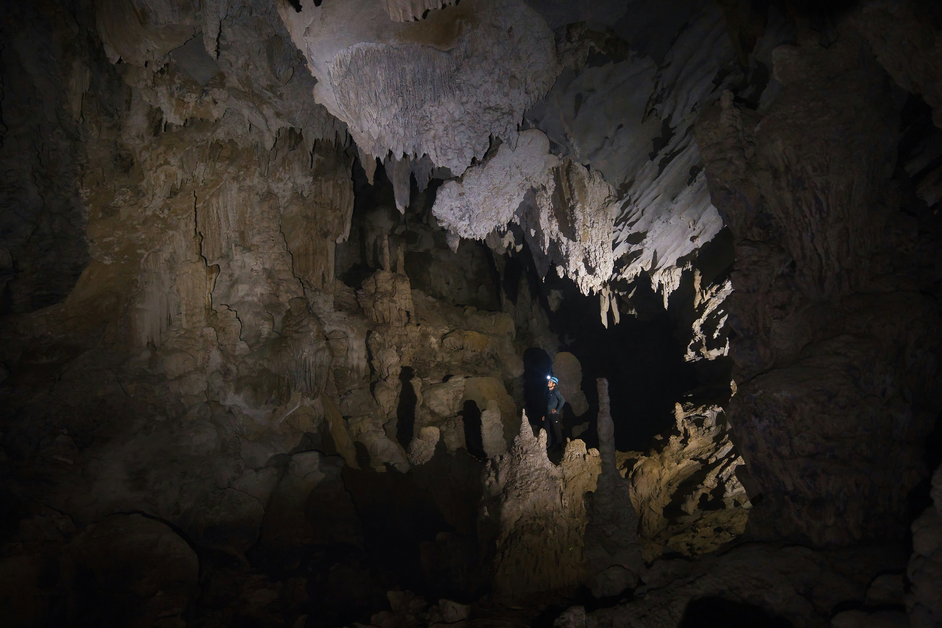 A person with a headlamp exploring a large limestone cave with hanging stalactites