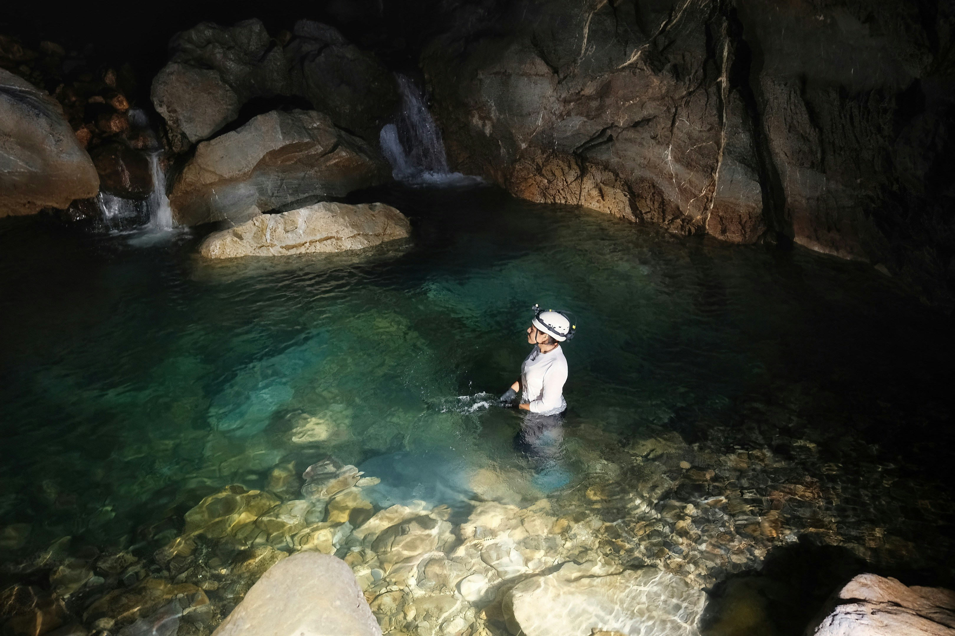 A person wearing a headlamp wading through a clear pool inside a dark cave
