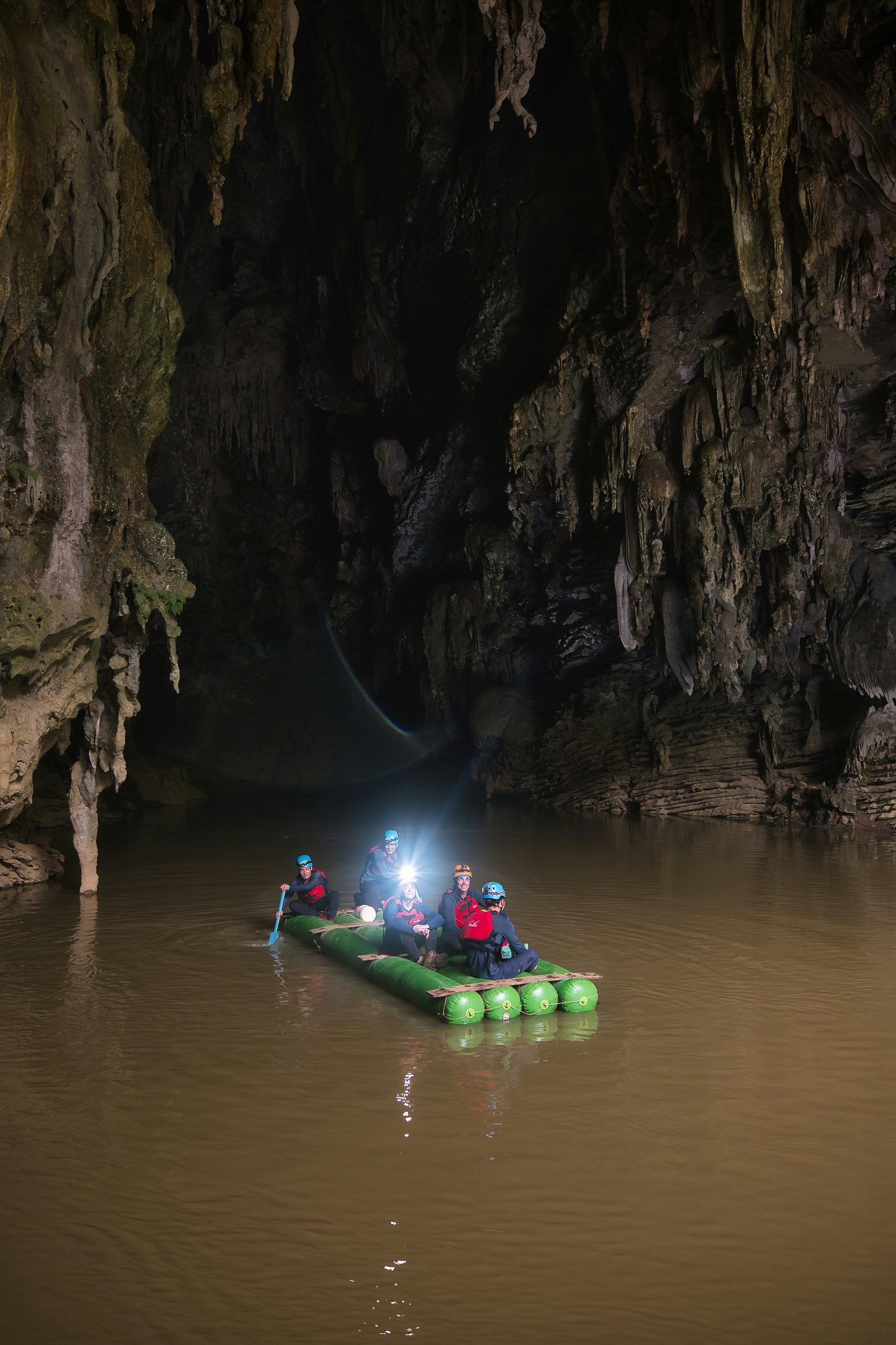 Five people on a green raft in a dark cave with stalactites
