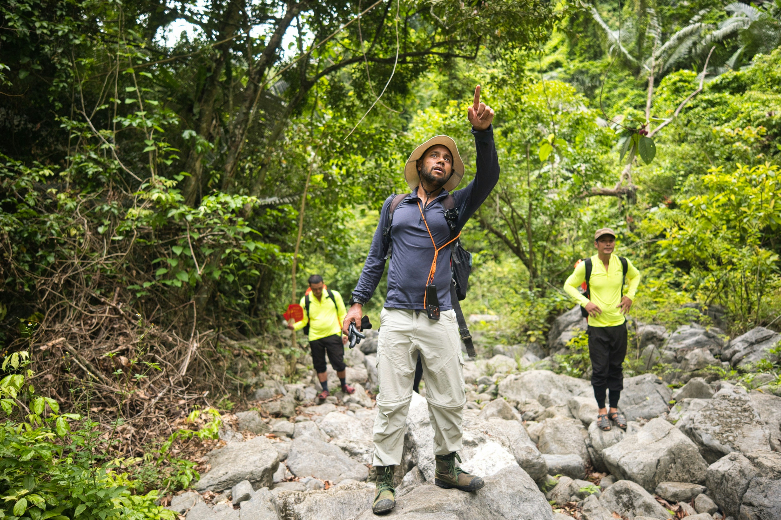 A person in a sun hat pointing upward on a rocky forest trail