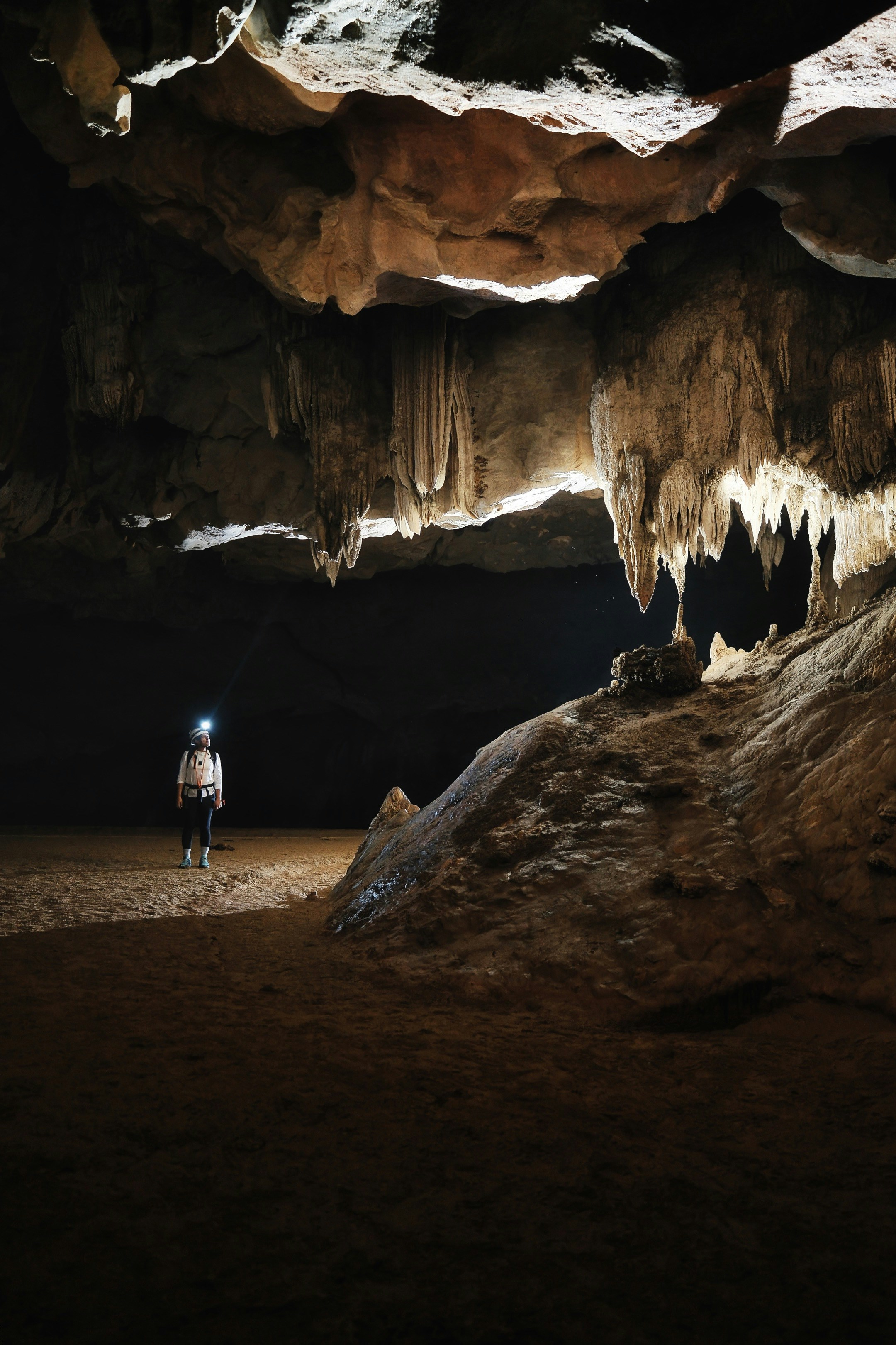 A person with a headlamp in a large cave with hanging limestone stalactites