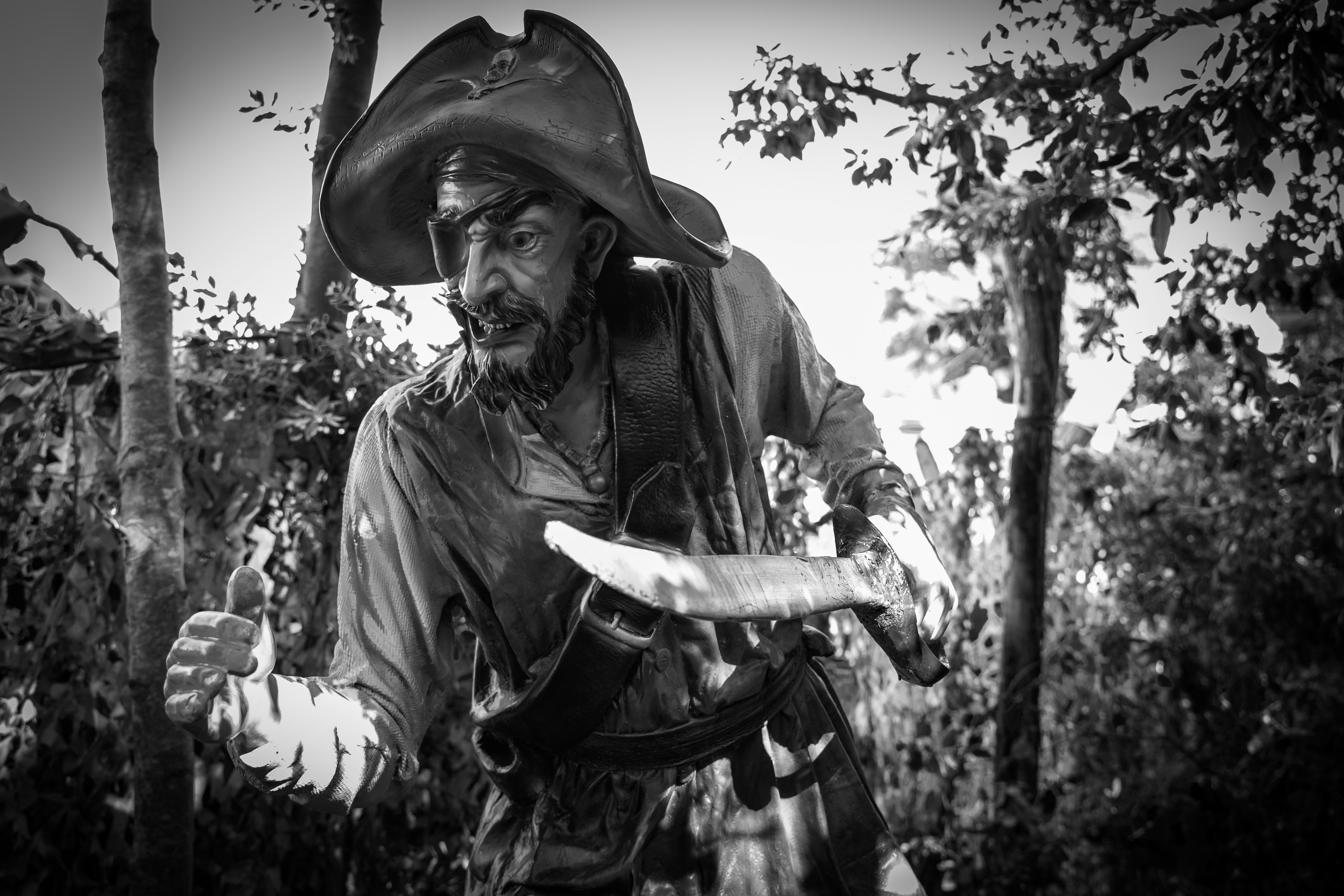 Dramatic black and white close-up portrait of a fierce pirate figure in full attire, wearing an eye patch and a tricorne hat, clutching a curved sword in a jungle-like setting.
