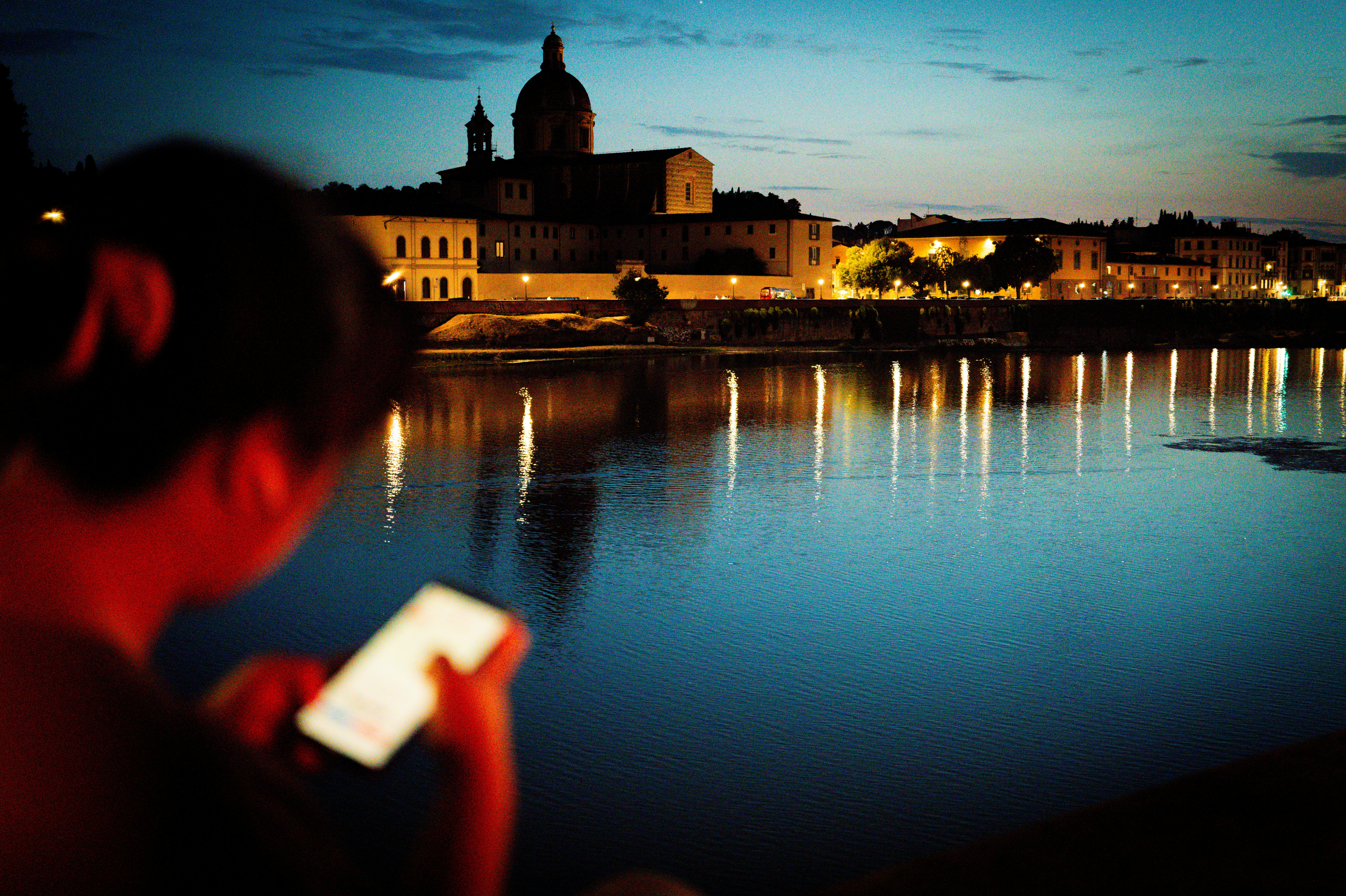 A person using a smartphone by a river at night with illuminated buildings.